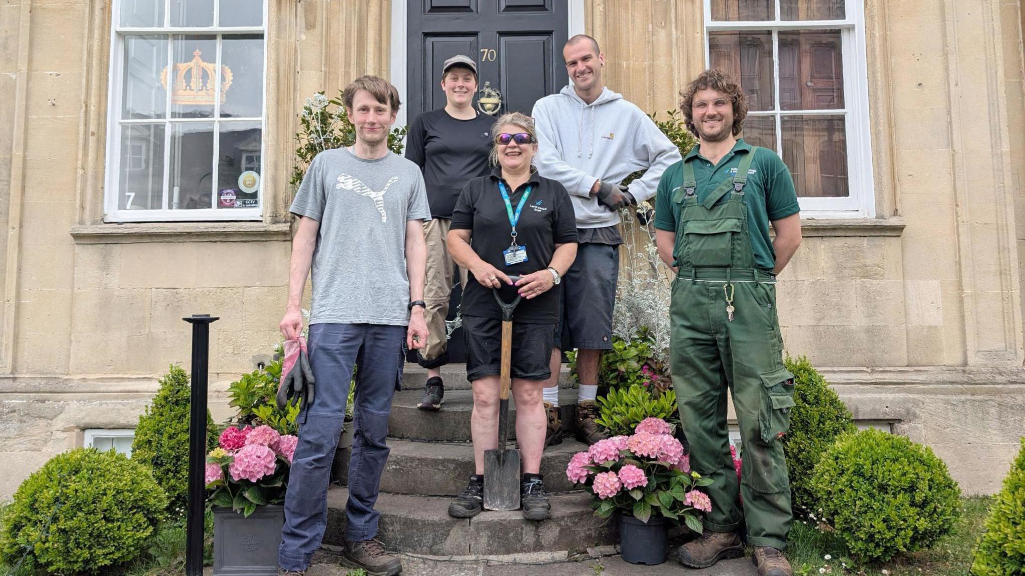 Five people stand in front of a Georgian-style house, on the steps leading up to it. There are flower pots and small hedges going upwards to it.