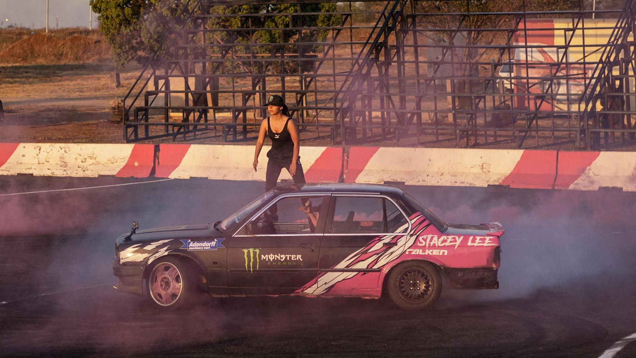 Stacey-Lee May stands on the windowsill of a black and pink car with her name on it as its locked wheels create smoke at a race track with bleachers in the background behind red and white crash barriers