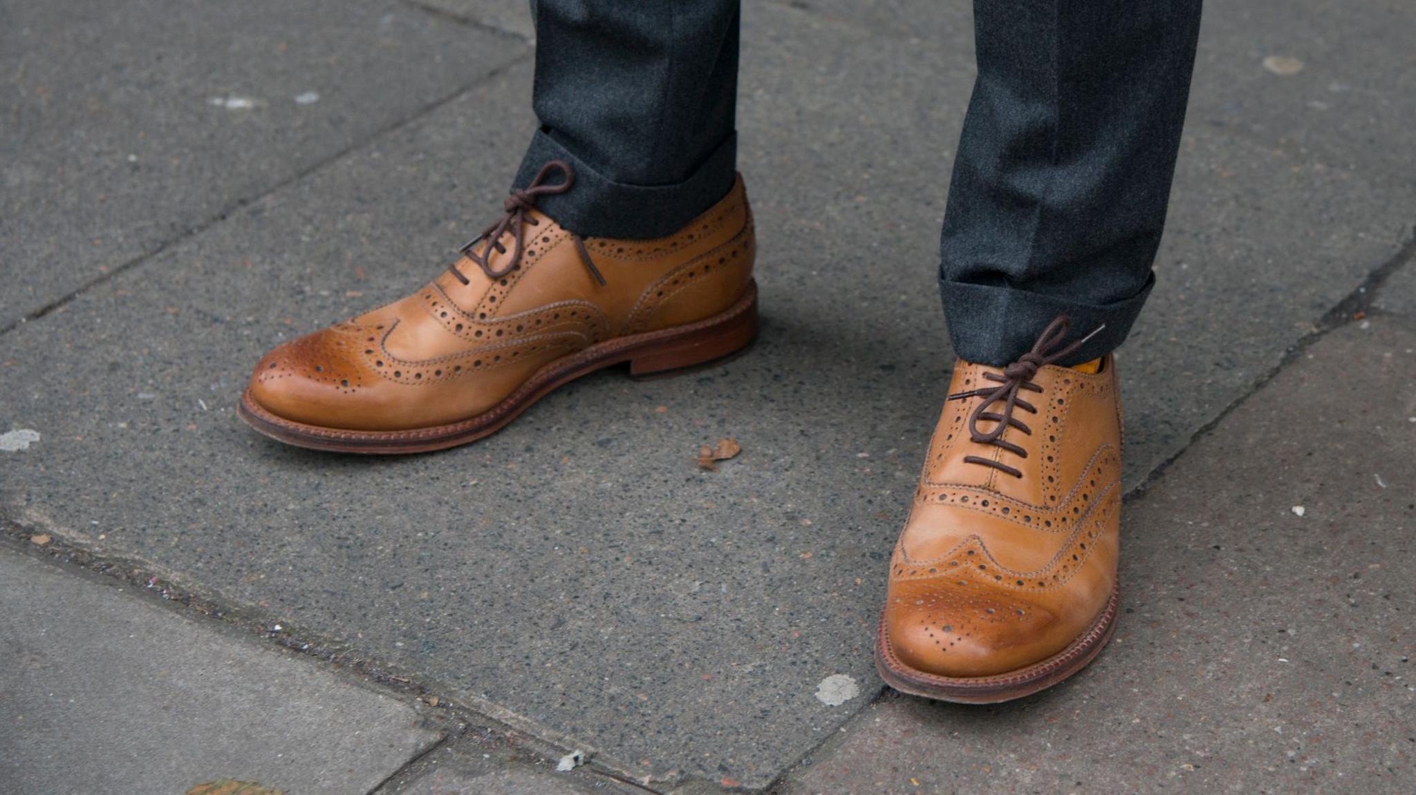 A close-up of a pair of brown brogue shoes by Grenson, on a man's feet, standing on a road. They are laced up and the man is wearing blue trousers.