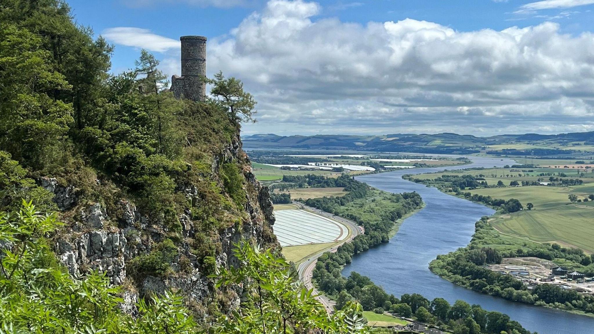 A view of a tower on a grassy embankment. To the right, down below, a river zig-zags through fields and bushes. It is a cloudy day with some blue skies peaking through.
