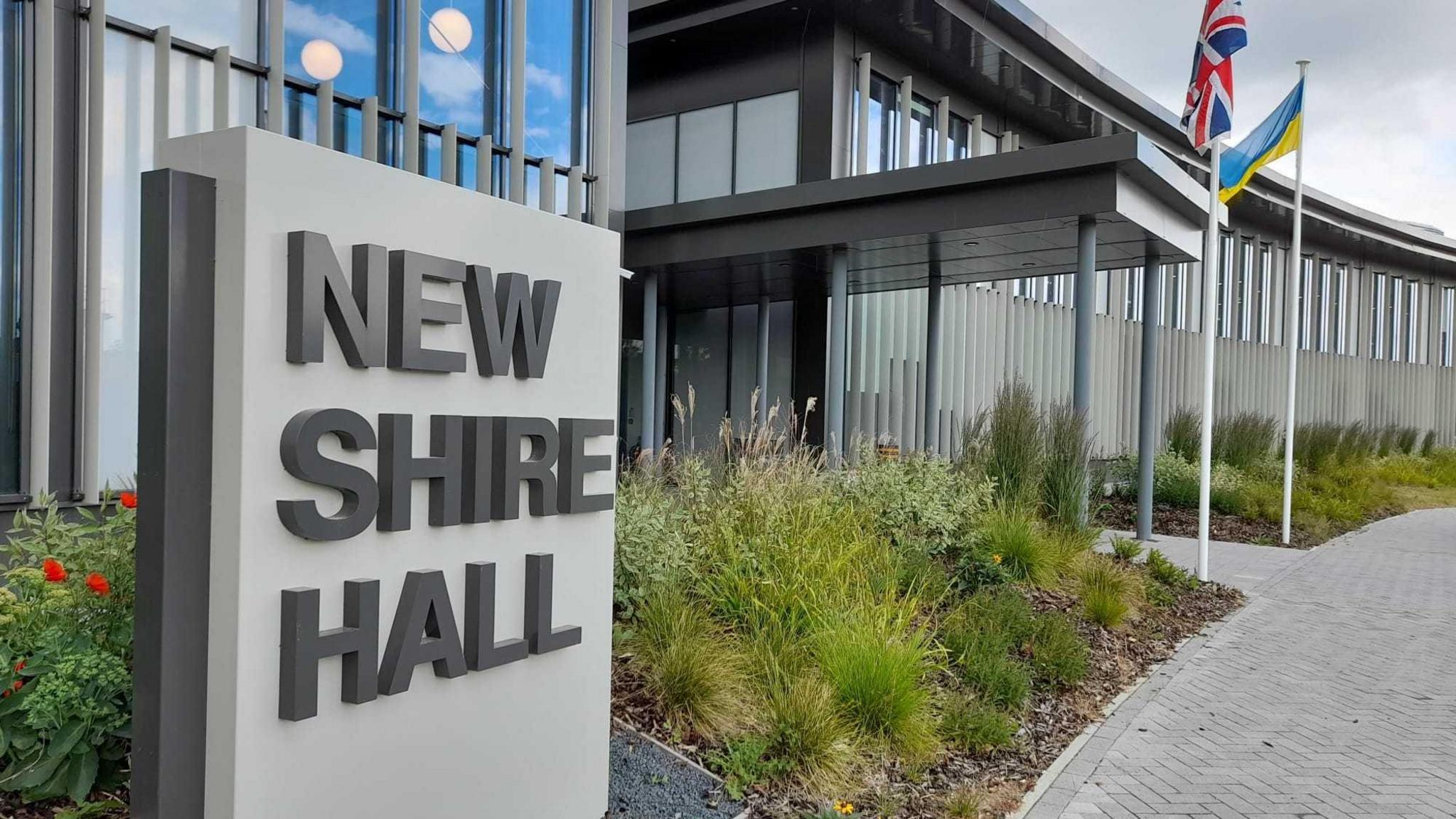 Exterior of council building with a sign saying New Shire Hall. Building is modern and there is planting of grasses and flowers around entrance. two flag- the Ukrainian and British are on poles. 