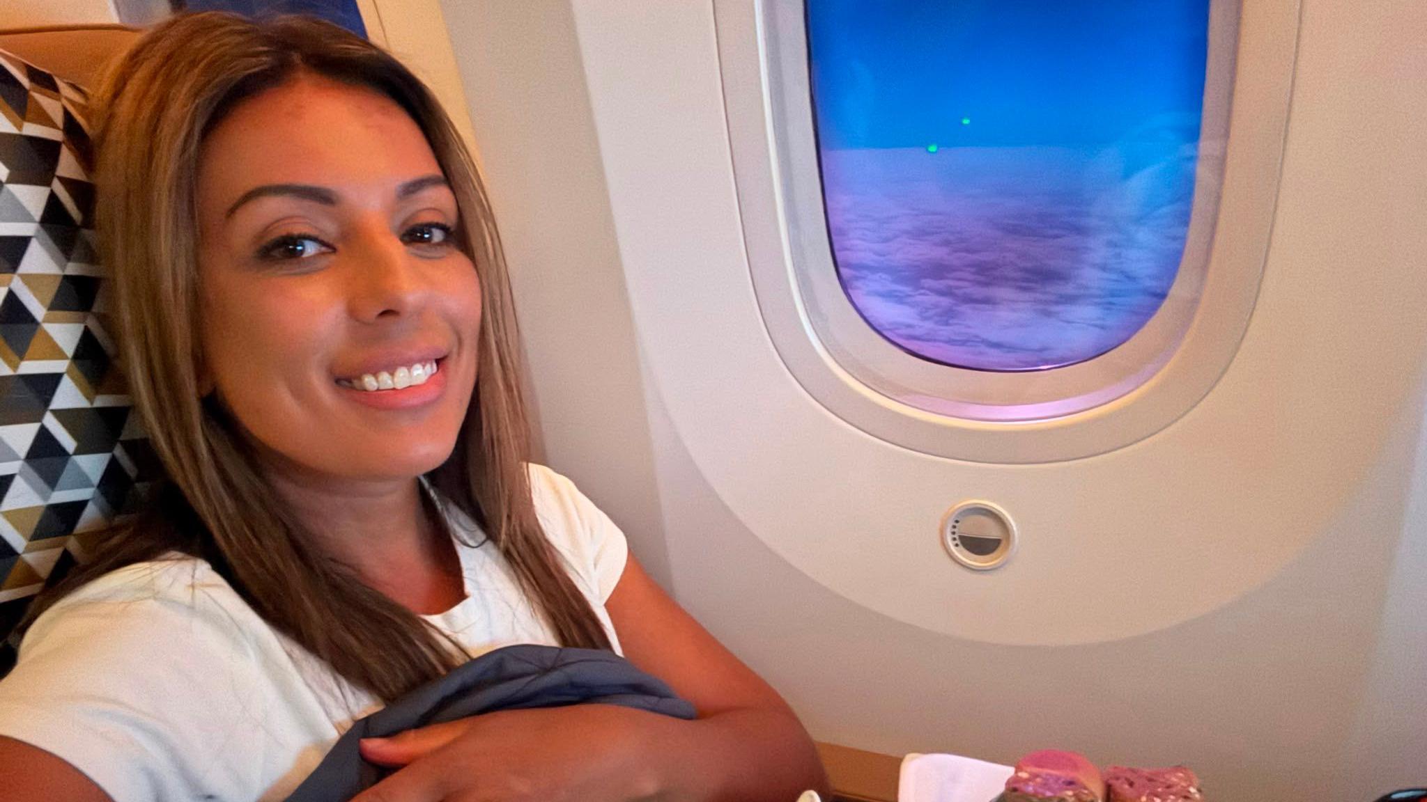 Karis smiles while sitting in an aircraft seat, a dusky blue sky with a thin layer of clouds outside the window.