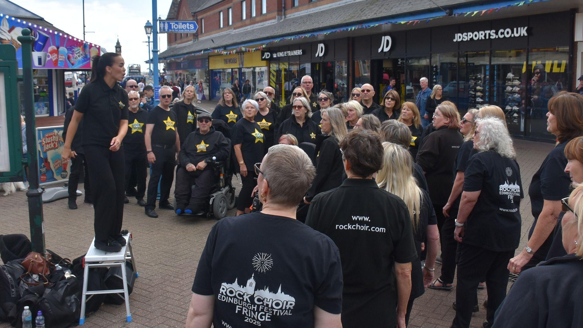 Lulu stands on a bench to conduct the choir members who are dressed in black t-shirts with Rock Choir on them. They have gold stars on their chests.