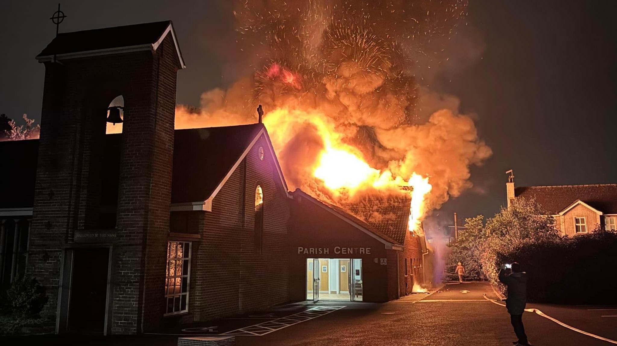 A night time photo showing the roof of the parish centre in flames, as a person standing taking a photo of the very well-developed fire