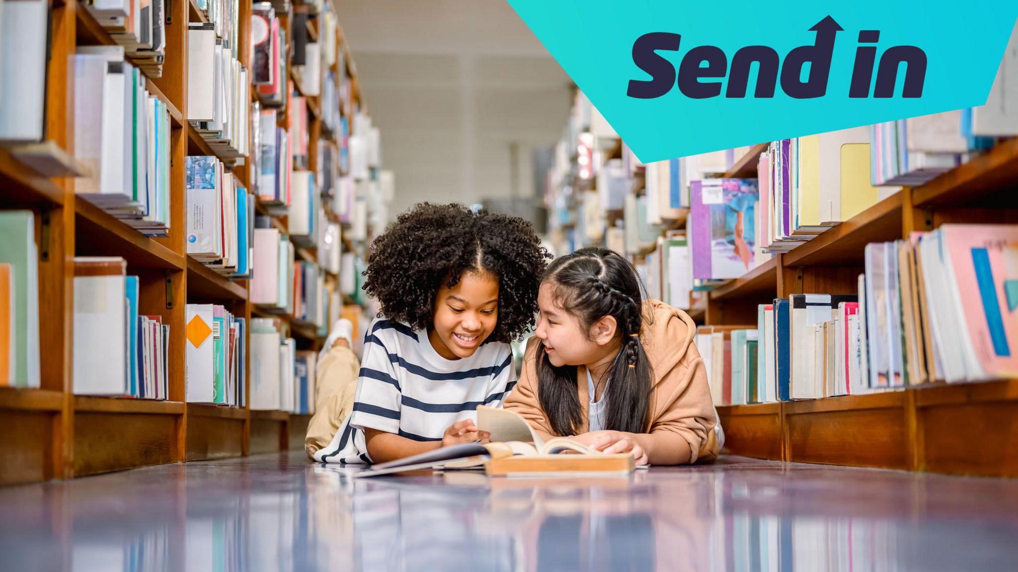 Two children sitting on the floor of a library reading a book