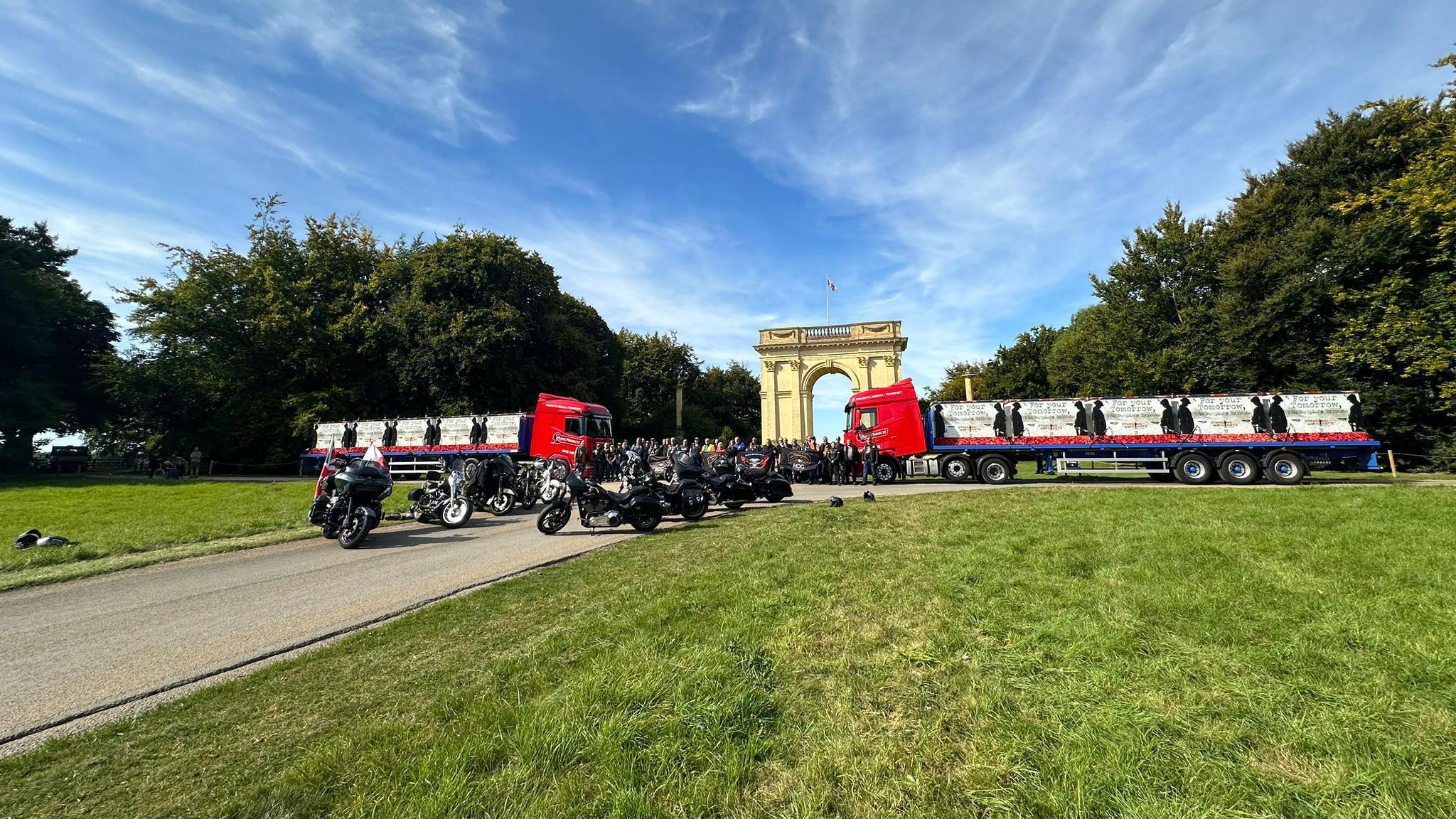 A convoy of two lorries arriving at the gates of Stowe Gardens. You can see a large number of motorbikes, green grass and a Grecian style archway. There is a path and plenty of trees.