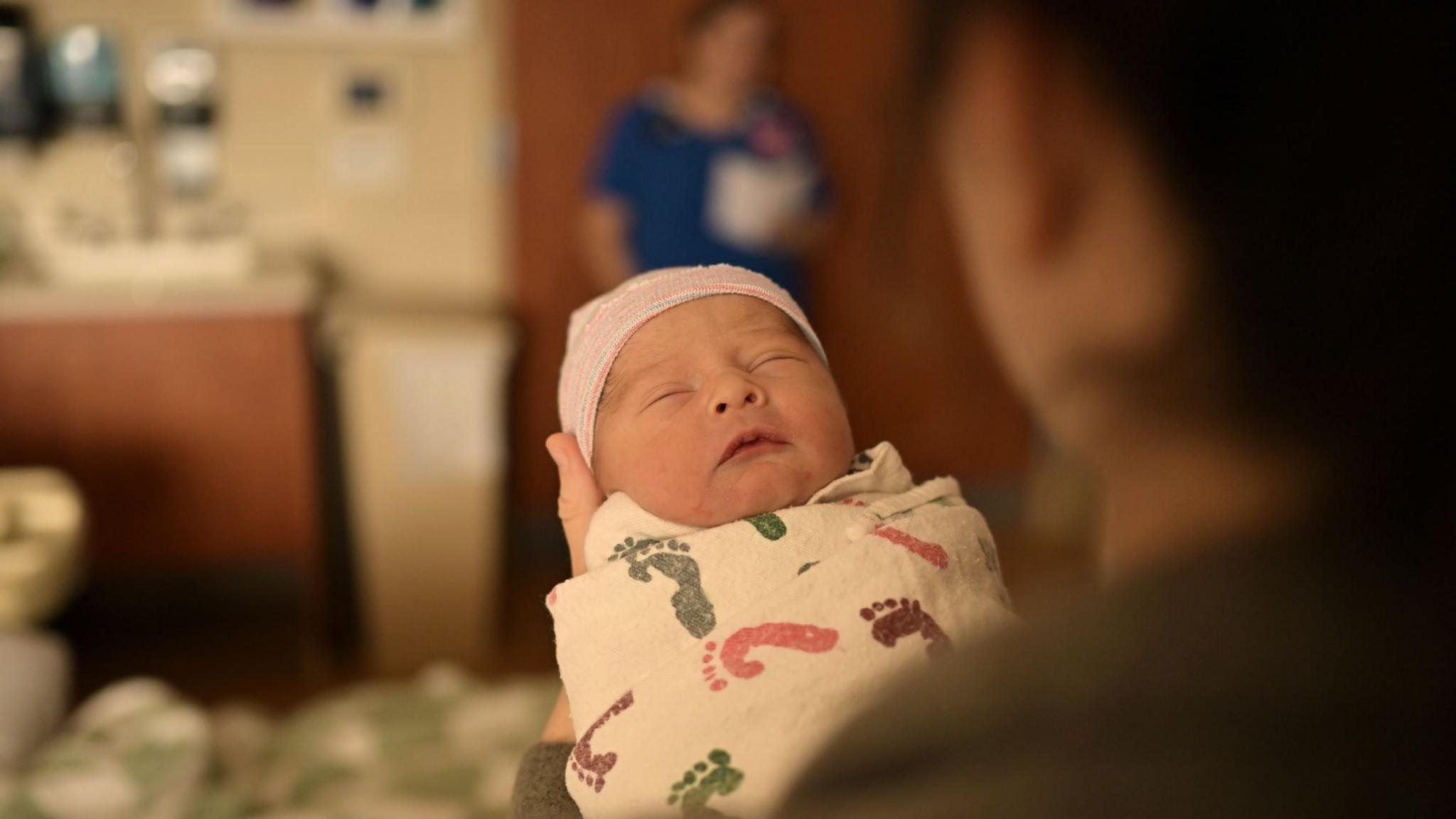 A newborn baby wrapped in a swaddle and wearing a pink hat being held by an adult in a hospital. A midwife stands in the background.