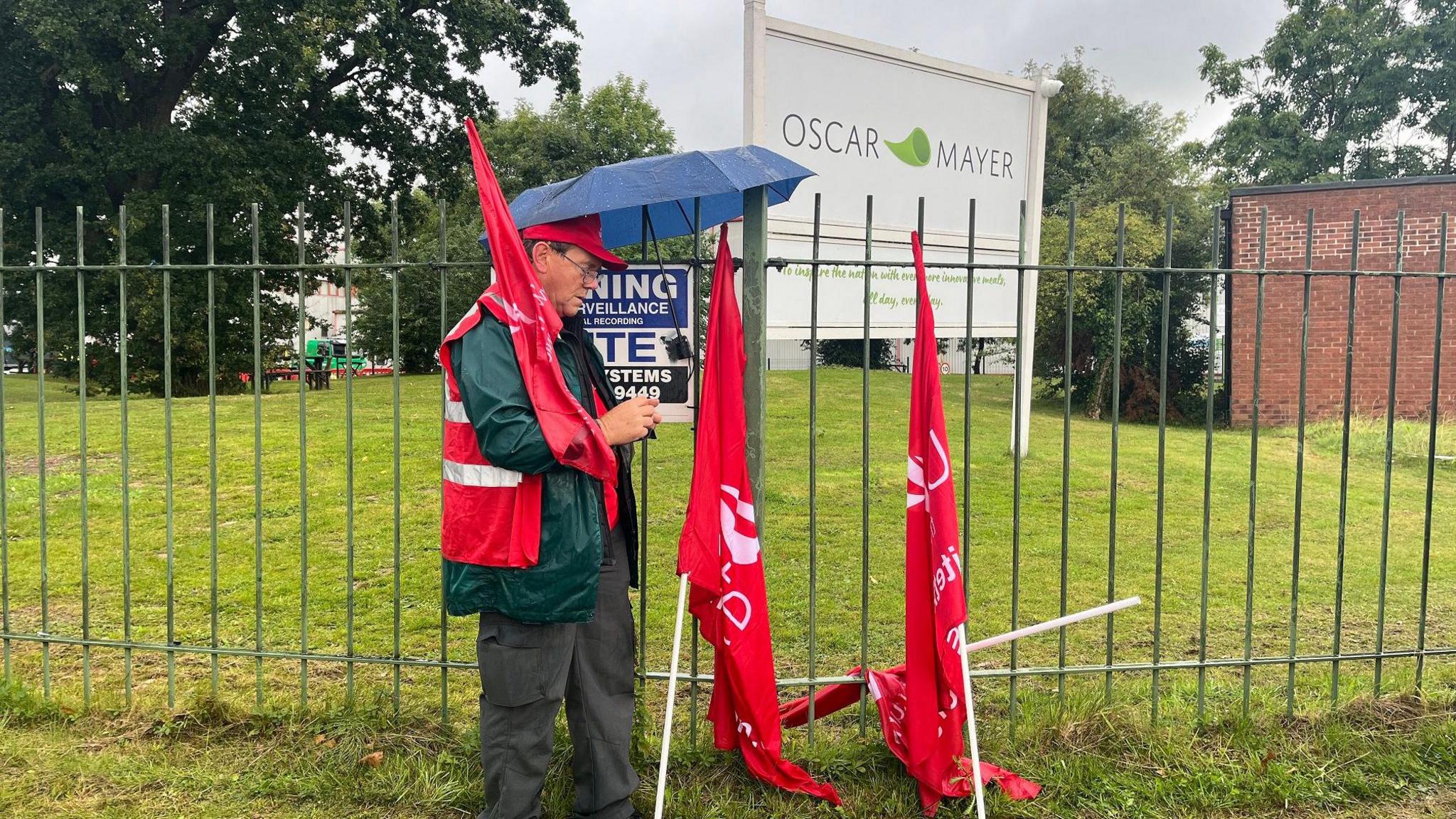 A Unite union representative, carrying red Unite flags in front of railings outside the factory, with the company sign in the background