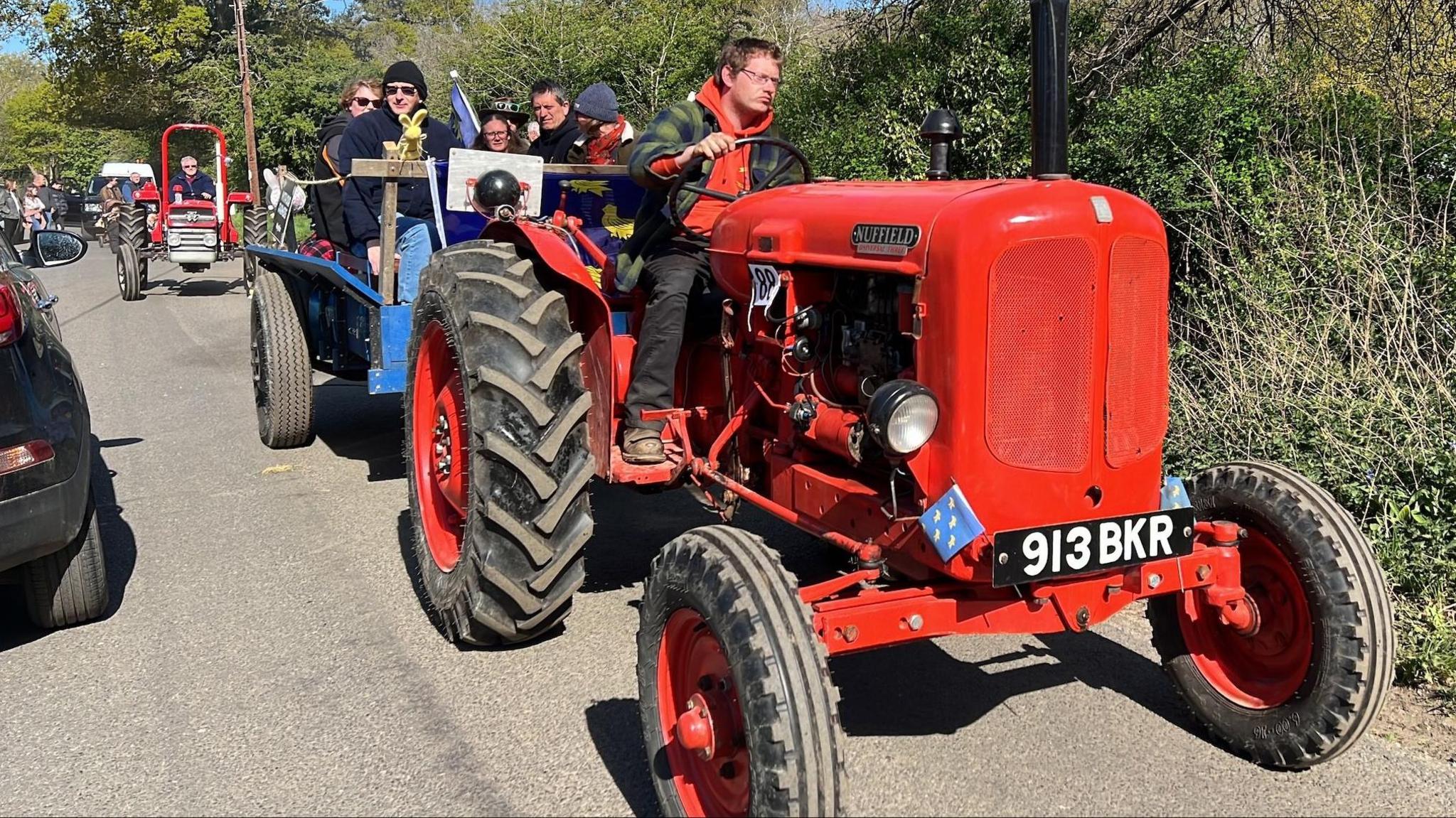 A man driving a small red tractor along a country road, with a blue trailer full of people in tow. There is another red tractor behind the trailer.
