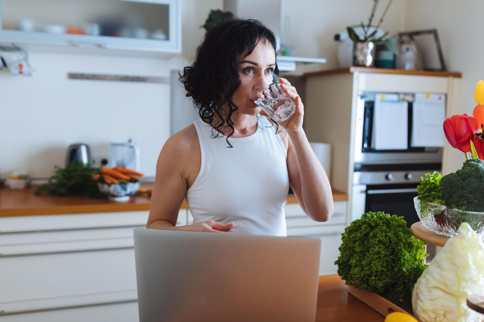A woman in a white top drinks water in a kitchen surrounded by fresh vegetables and a laptop