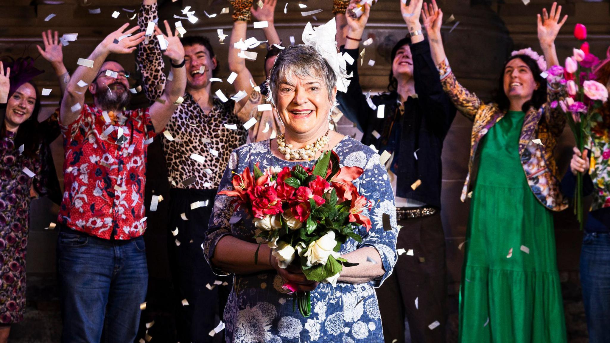 Image of Allison Gardner - a woman with short hair, wearing a blue dress with white floral print, holding a bouquet of red flowers as she is showered with white confetti by a group standing behind her wearing wedding guest type outfits, styled to the 1980s - bright prints and animal prints. All are smiling and celebrating with white balloons at their feet and waving their hands in the air.