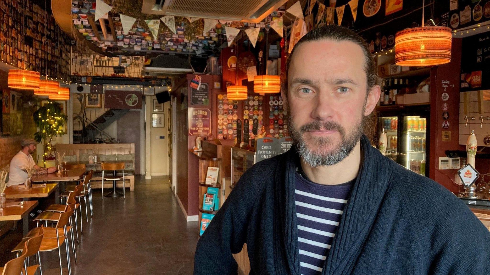 A bearded man with a scraped-back ponytail wearing a blue cardigan and blue and white Breton top underneath standing in a bar with a man sitting at a table in the background. 