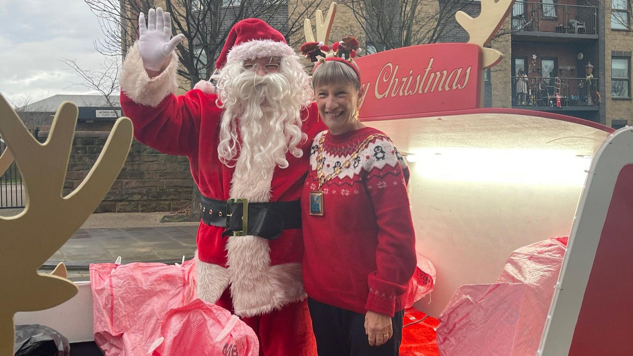 Councillor June Burns stood on a Christmas sledge with father Christmas. She has short hair and is wearing a red Christmas jumper and black trousers.