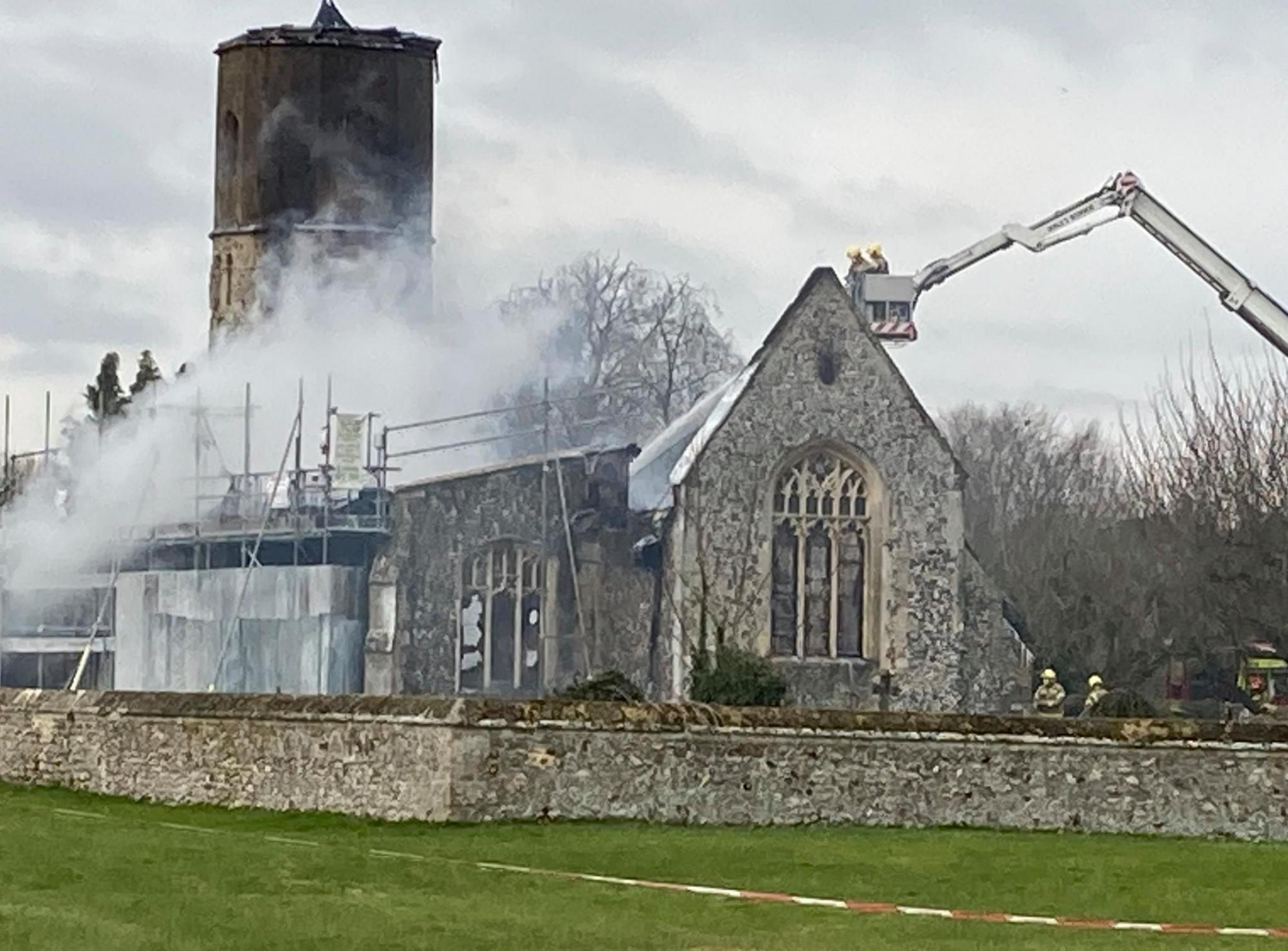 The remains of the church with a fire service platform ladder hovering over it. One side of the building has scaffolding on it covered in metal hoardings. The roof has disappeared.