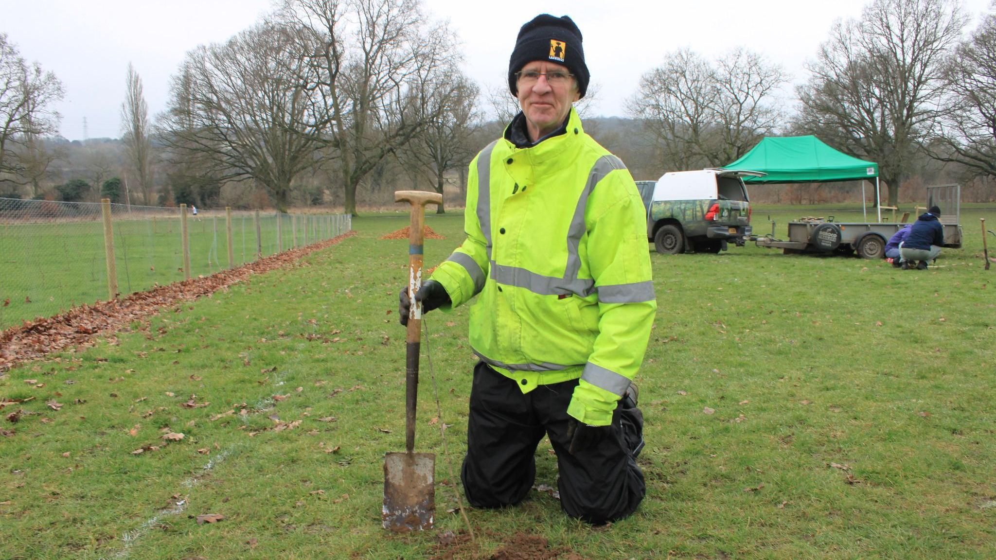 A man wearing a high-vis jacket on his knees holding a shovel. He is looking into the camera