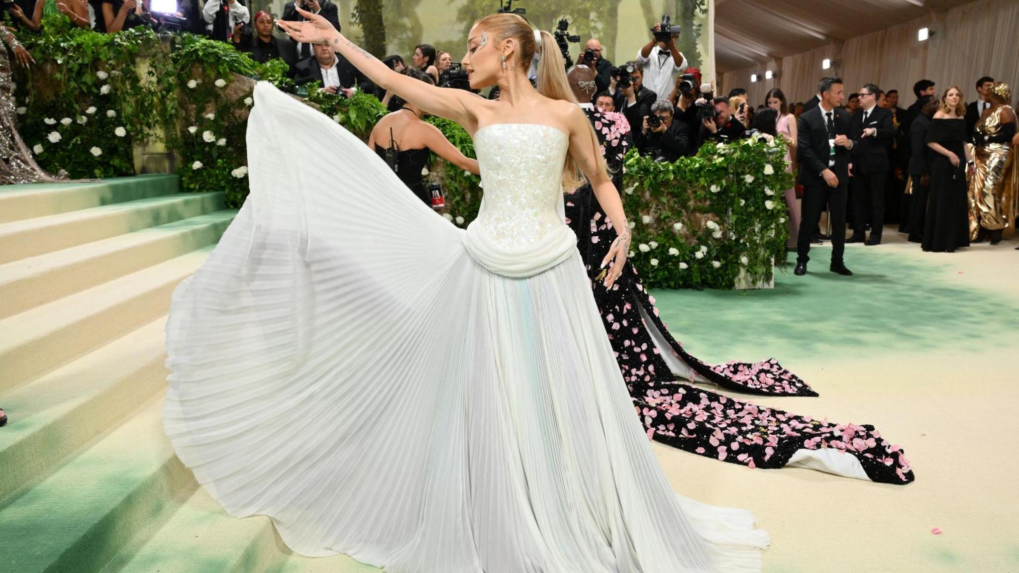Singer Ariana Grande is on the red carpet at the Met Gala 2024 in New York. She is wearing a floor-length, white ballgown and is showing off the pleats on the bottom. She has her head turned to the side and her long, blonde hair is tied back in a ponytail.