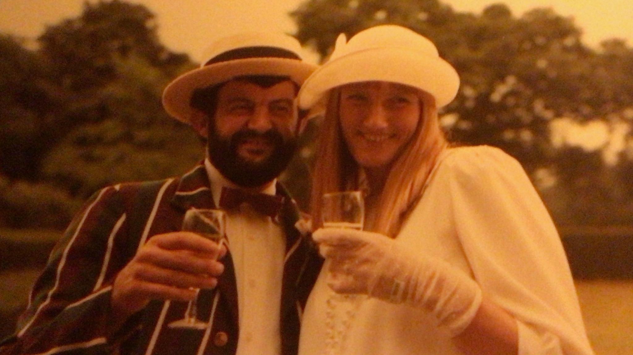 A man in a green and purple striped blazer and boater and a woman in a white coat and hat. Both have glasses of champagne in hand