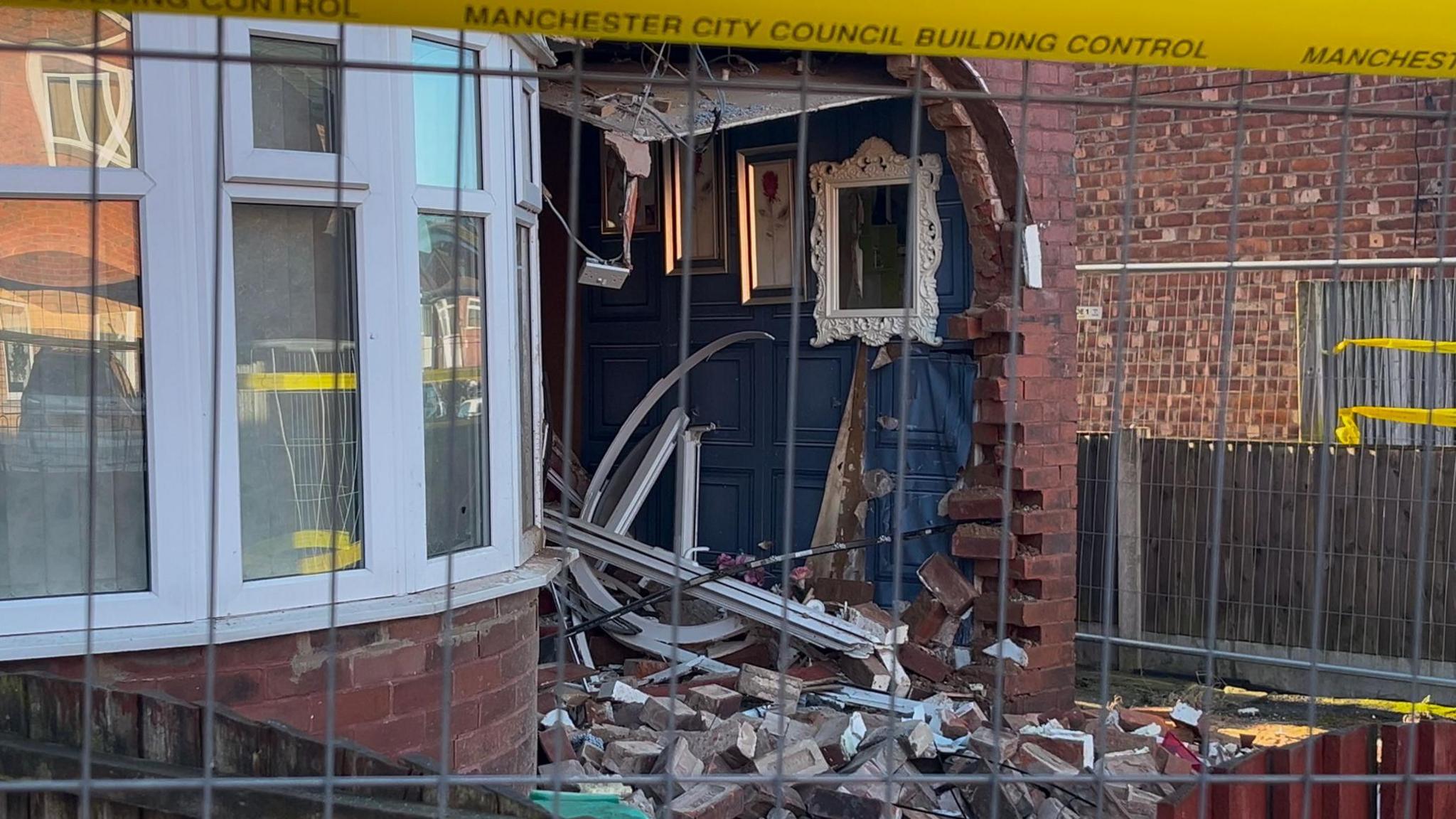 Close-up image of brick damage to the house's front entrance and interior. Paintings and a mirror still remain intact on the wall of the hallway.