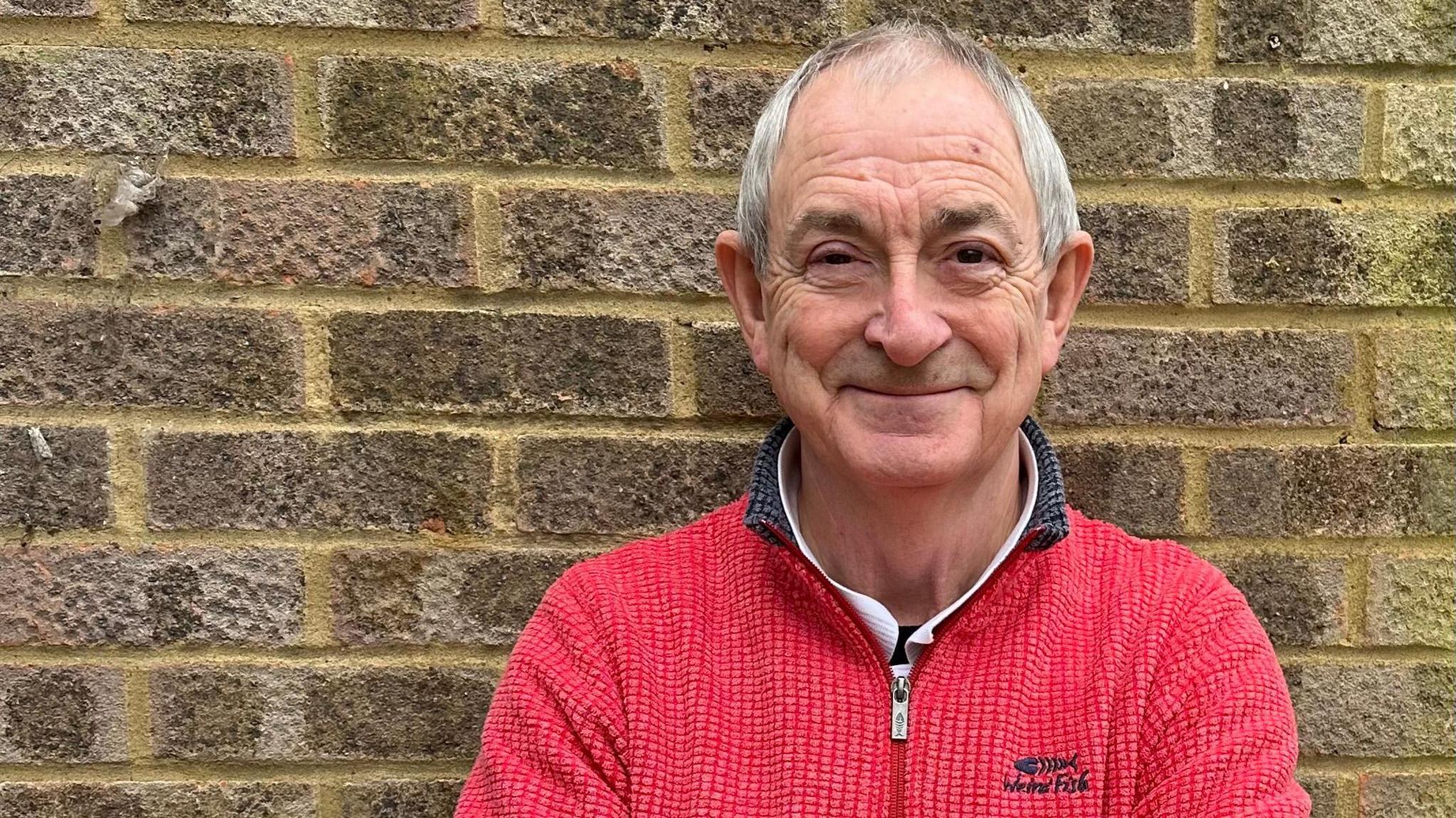 A man with grey hair and wearing a red fleece, smiling at the camera with a brick wall behind him.