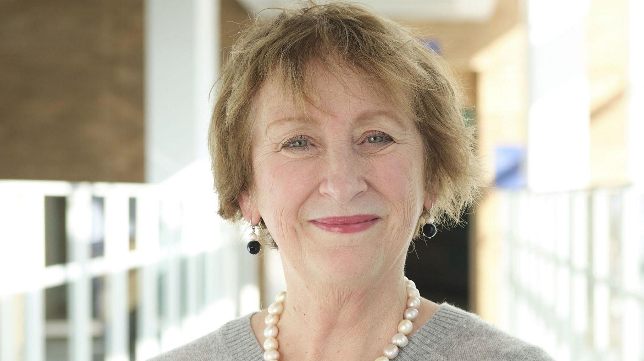 Lesley Dwyer smiling for the camera. She is wearing a necklace and a grey top. She is in the atrium of the Norfolk and Norwich Hospital.