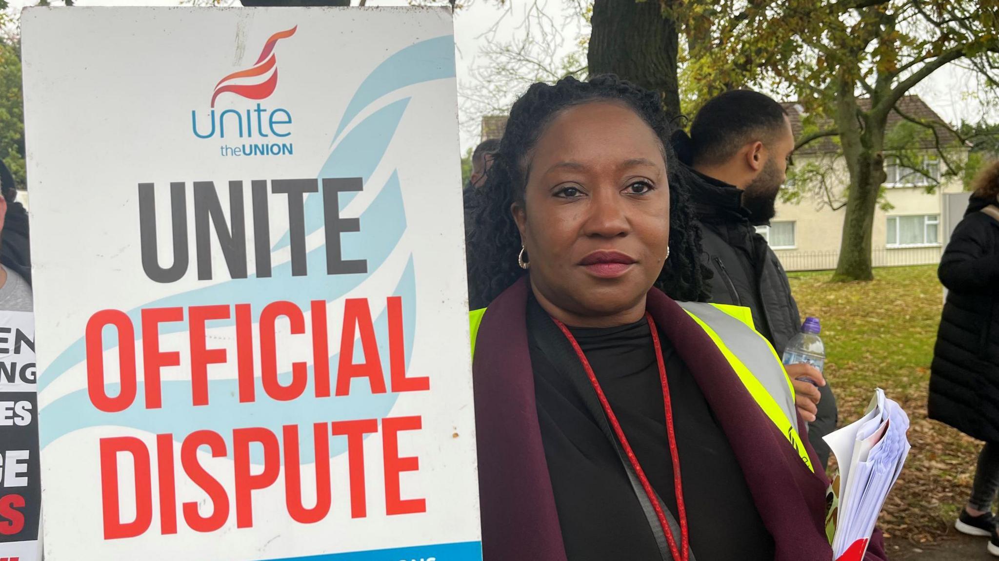 Sharon Harding holding a placard, wearing a purple jacket with a fluorescent vest and a red lanyard 