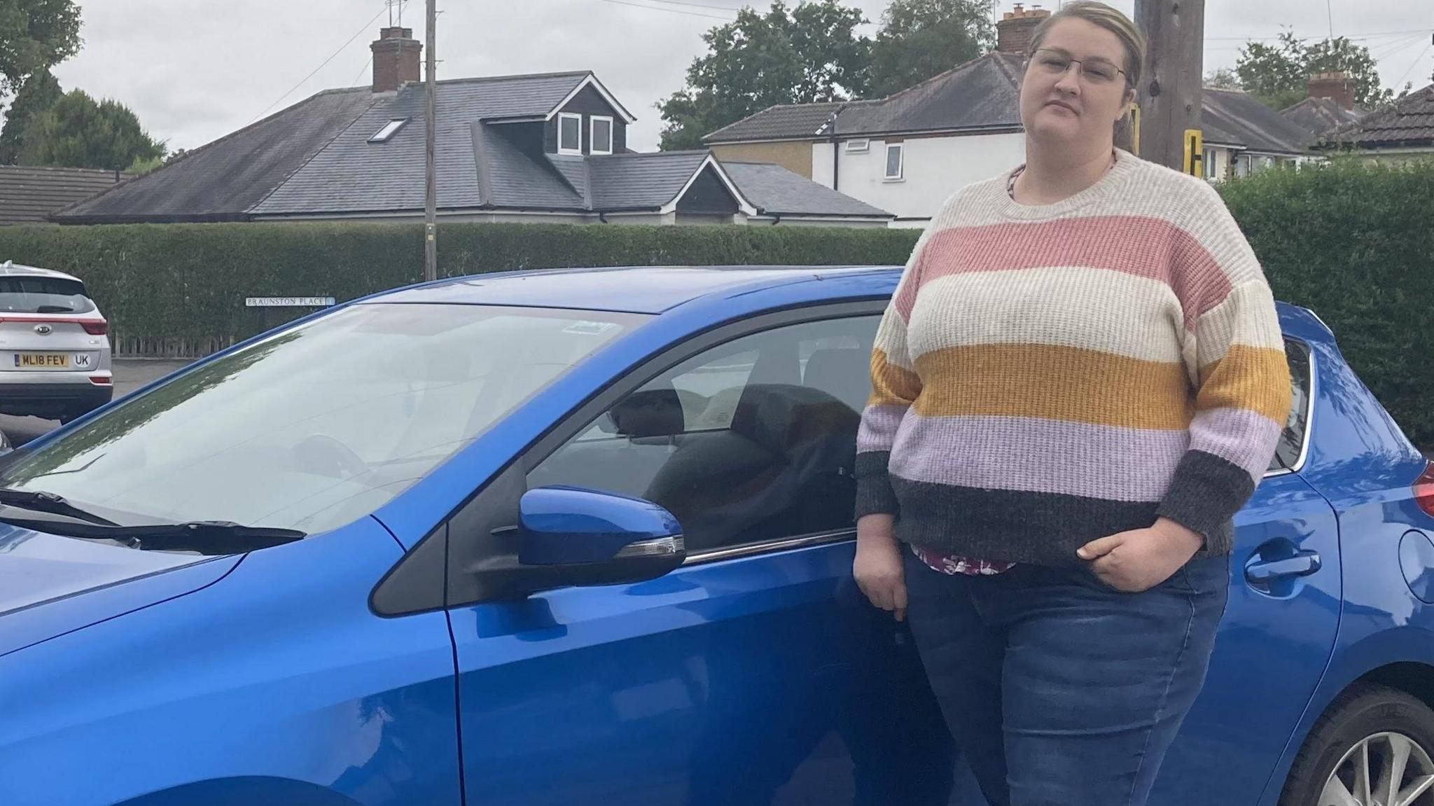 Mary Bates is standing in front of her blue car outside her home.