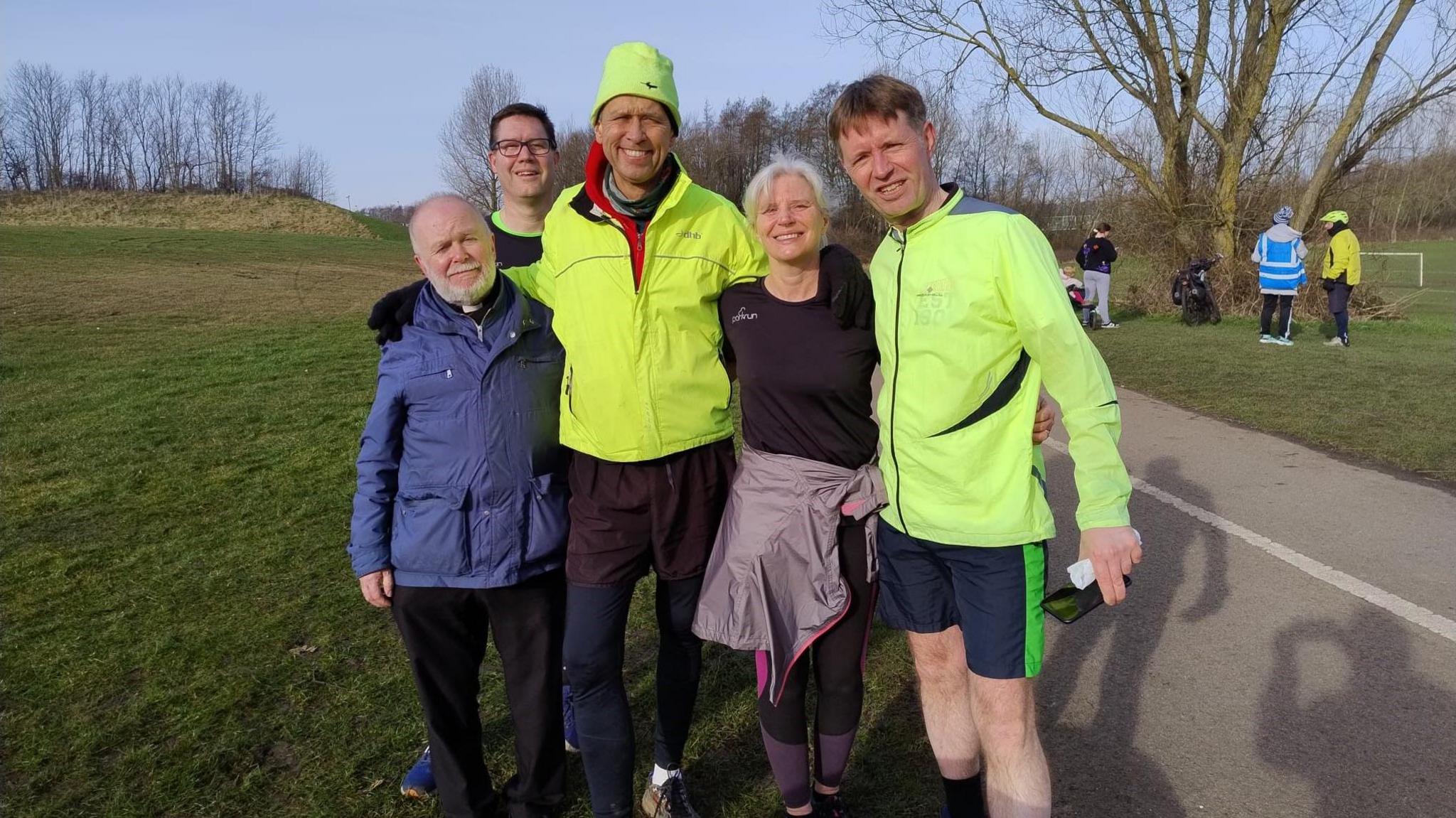 A group of four male and one female vicars, including David Chadwick. Four are wearing sports clothing including hi-vis waterproof jackets. One is wearing a thicker, blue winter coat. They are standing in a park on a grass area next to a walking and cycle path. A few other people can be seen behind them and there is a football pitch in the distance.