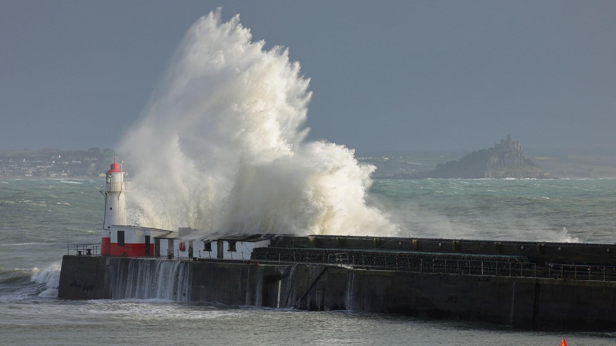 A huge wave hits the quay. It towers above the lighthouse. St Michael's Mount is visible across the bay to the right and part of Penzance can be seen to the left in the distance.