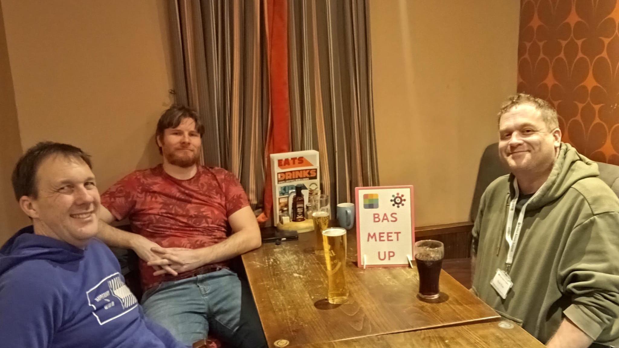 Three men sit around a pub table with drinks. They are looking at the camera. There is a sign reading BAS Meet Up on the table. 