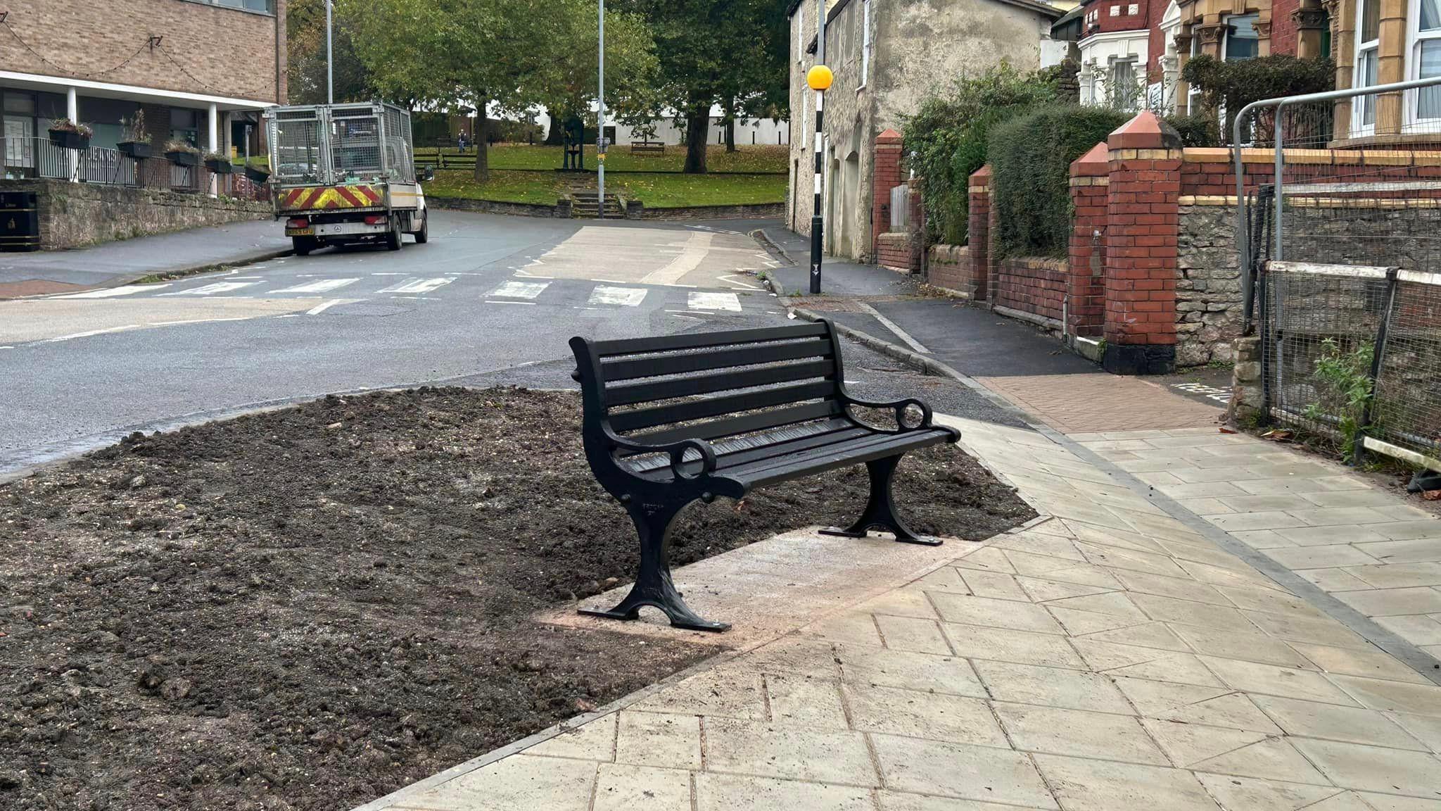 A new black bench installed on the side of the road, beside a freshly laid pedestrianised area of soil. In the background you can see a zebra crossing, a small park area and houses lining the street. 