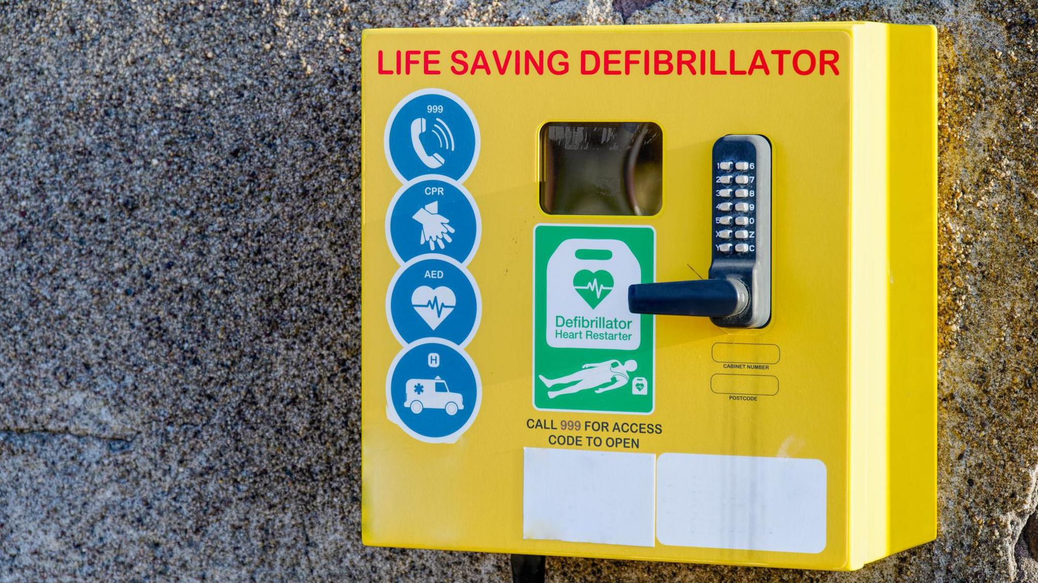 Yellow defibrillator box attached to pebble-dashed wall. It reads "life saving defibrillator" and has a code lock.