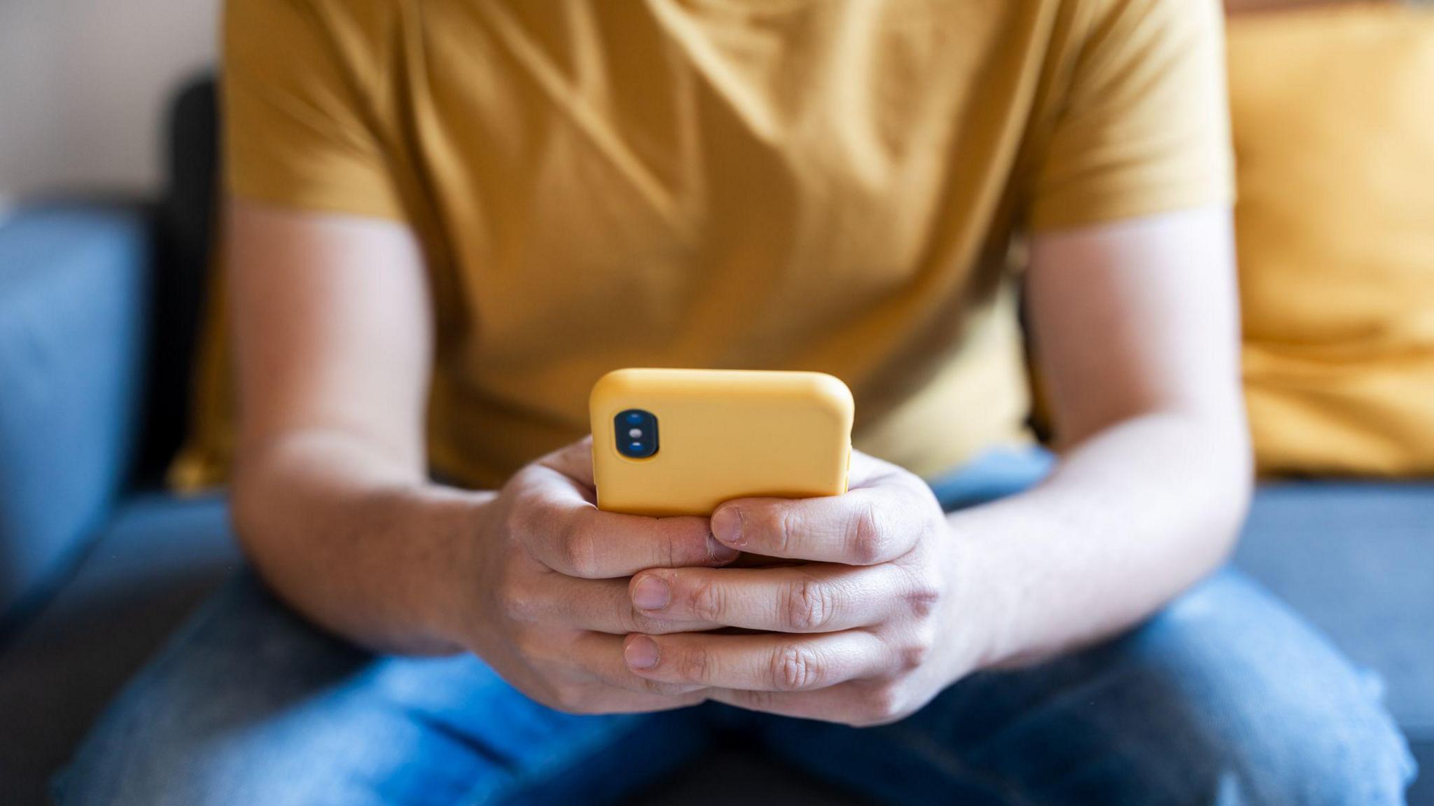 A man sitting on a sofa wearing a yellow t-shirt and jeans, holding a yellow smartphone in his hands in front of him.