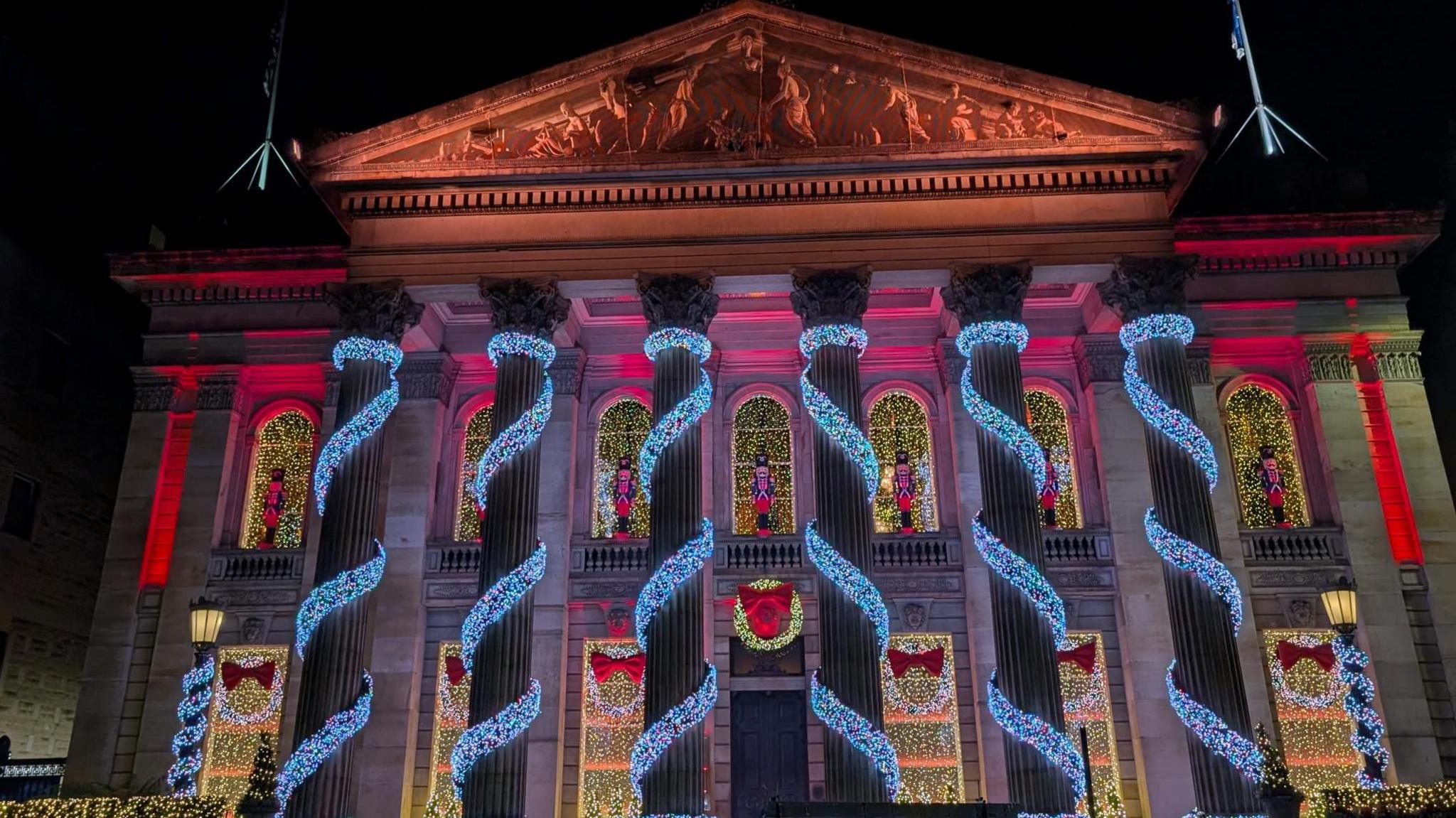 Colourful lights wrap around the six stone pillars at the front of the building. The windows behind are illuminated.