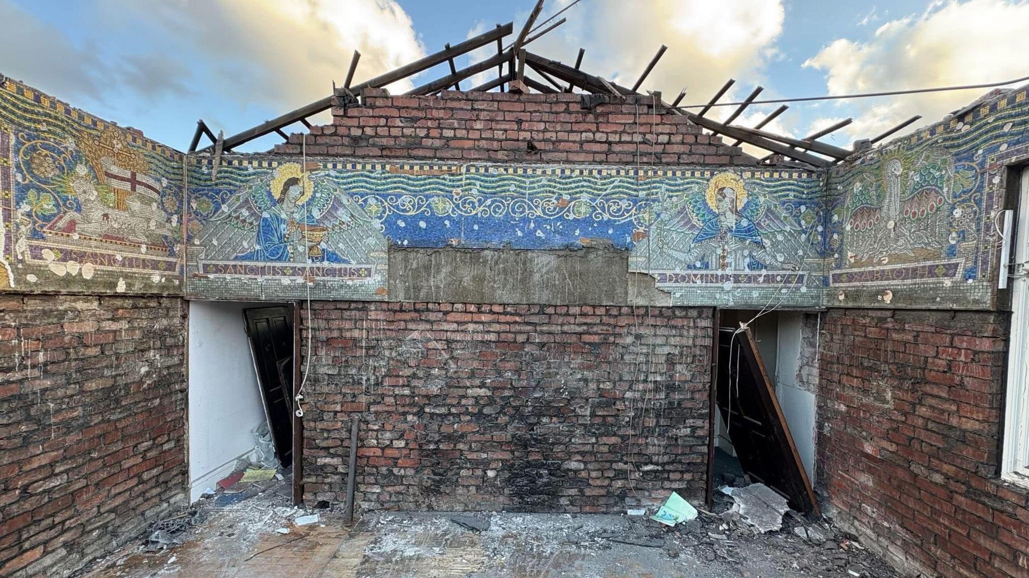 The exposed gable end of one of the convent buildings, which has been partially demolished, and has exposed a mosaic of two angels facing each other from opposite sides of the building.