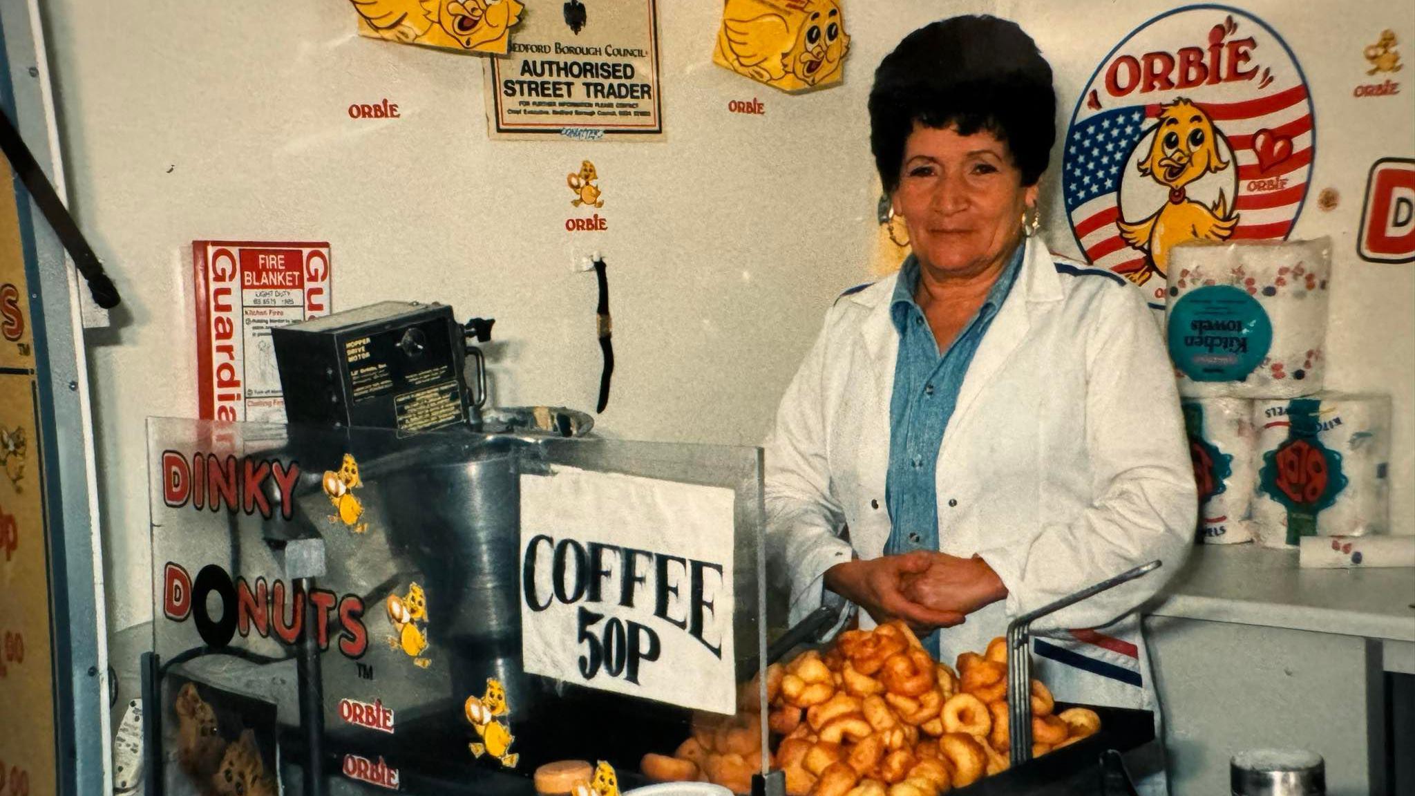 Lina Ognissanti selling donuts from her stall in Bedford several years ago. She is wearing a white coat and looking at the camera. At the front of her stall is a pile of freshy cooked small donuts and a sign advertising coffee.