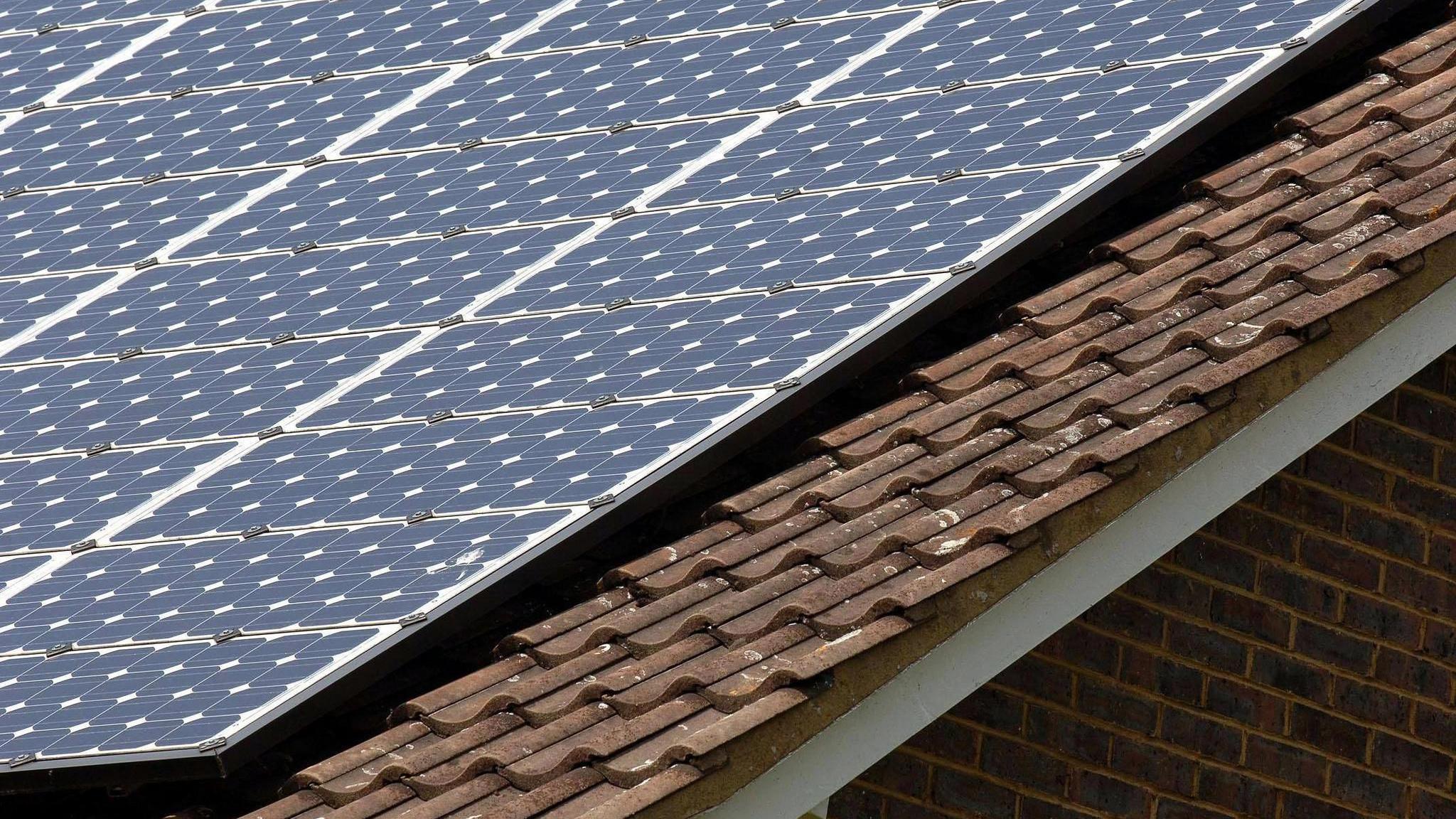 A sloped roof on top of a house which has fitted grid-patterned solar panels on it. It is placed above brown tiles on what looks like a sunny day.
