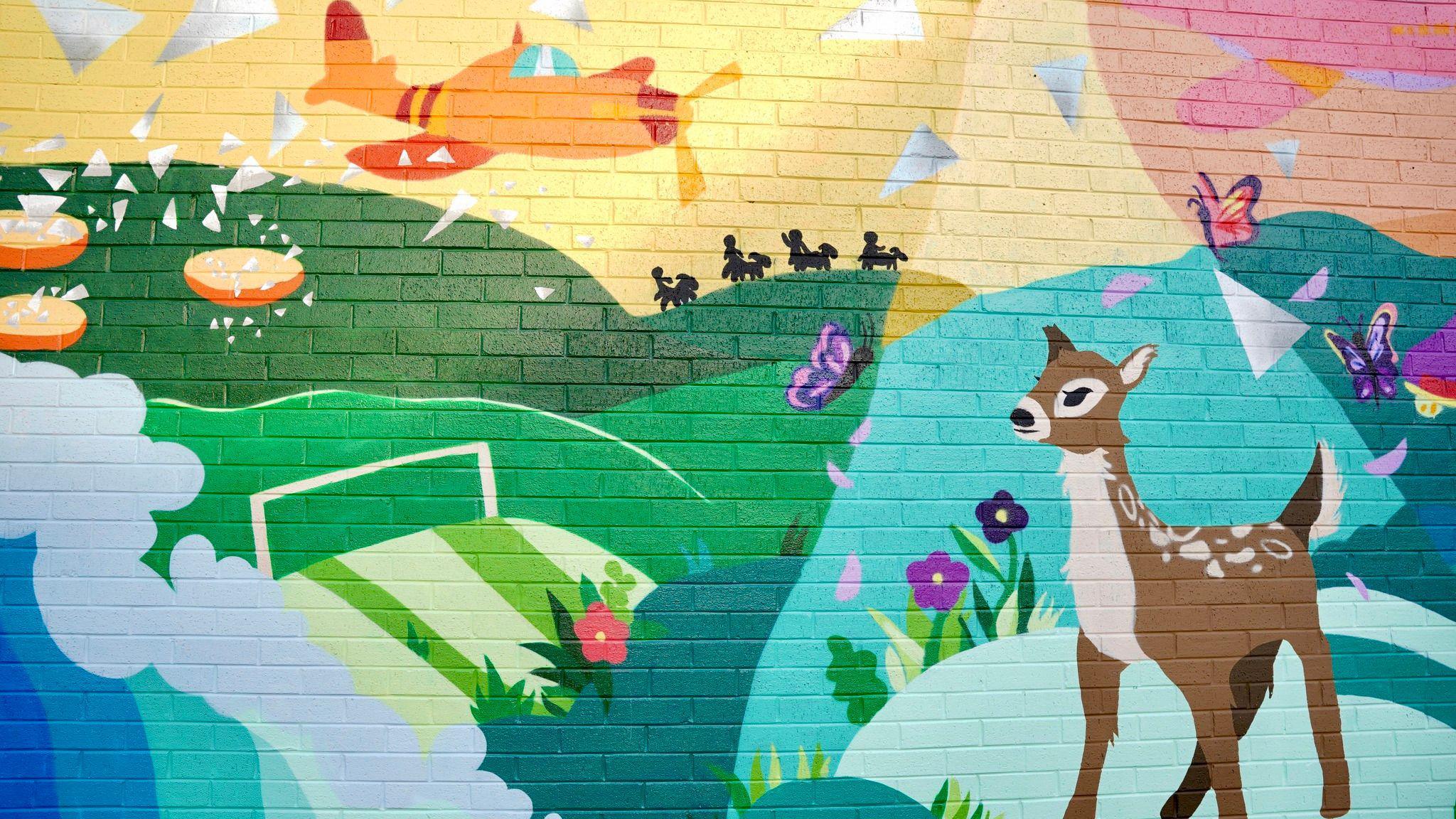 Part of a mural on a brick wall. It has rolling hills that are green, with splashing blue waves below it. There are purple butterflies flying and a brown deer in the foreground. The sky is pink and yellow and there is an orange plane flying across it.