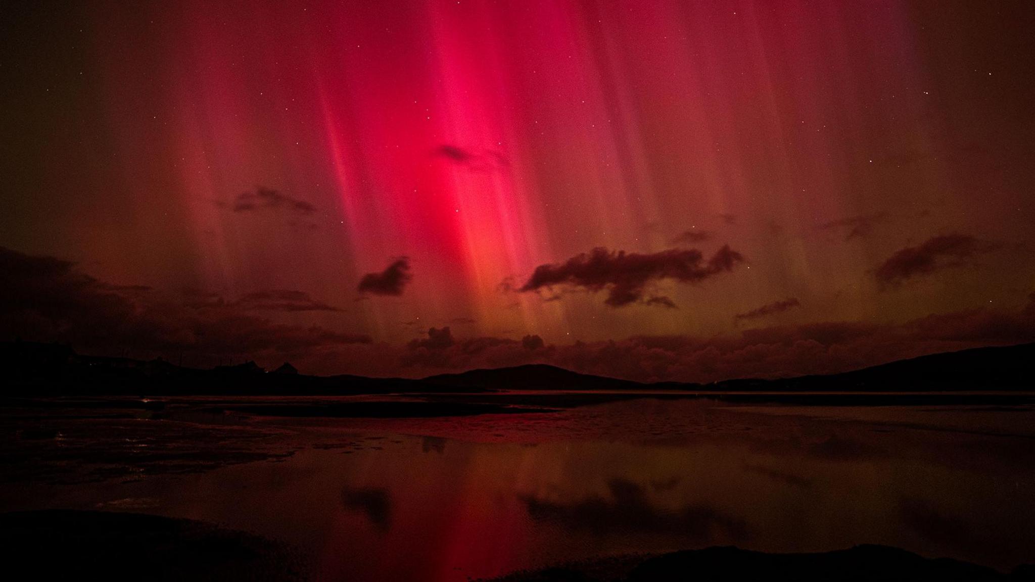 The still sea and a hill can be seen in the foreground, the hill silhoutted against some very deep red aurora.