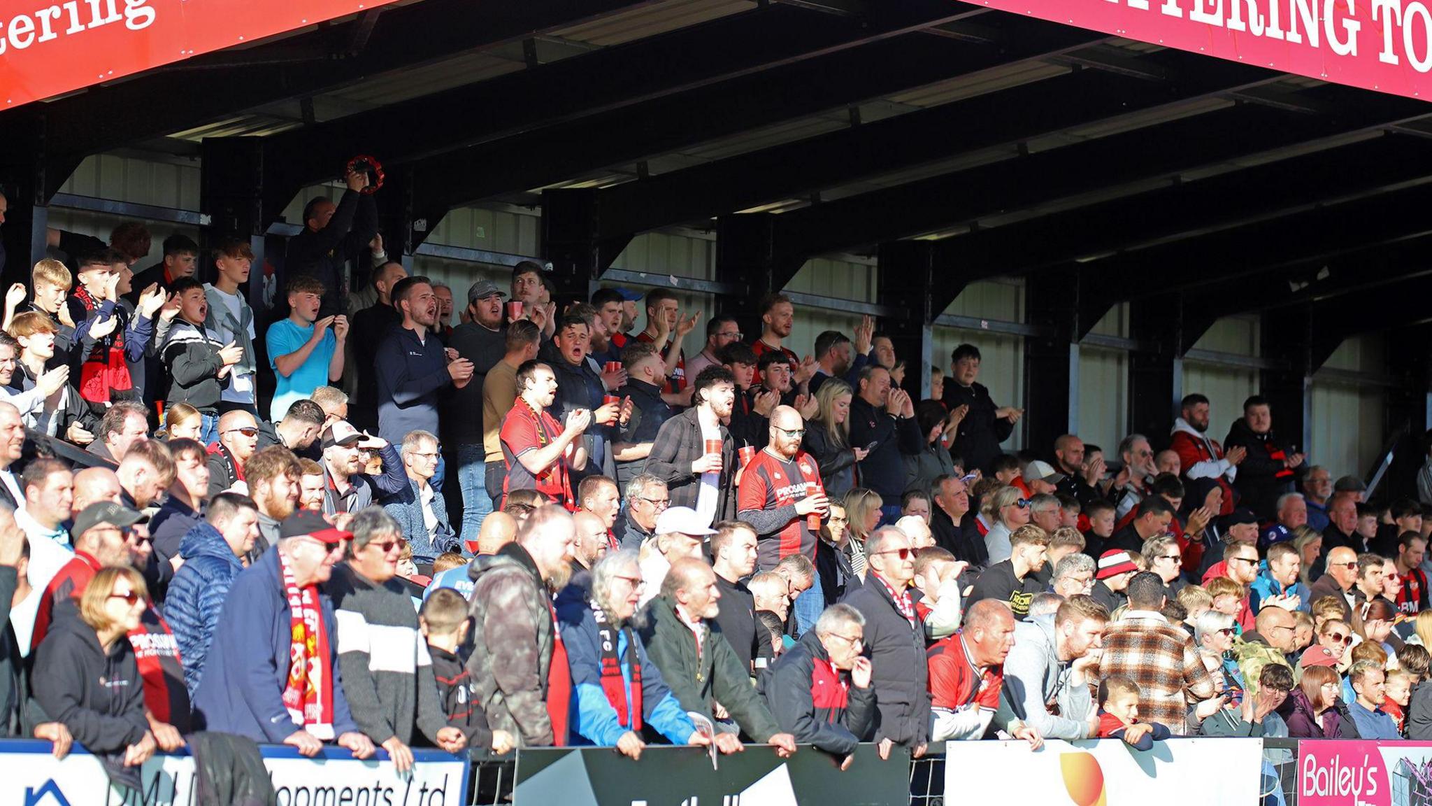 Kettering Town fans in a packed, terraced stand watching a game. The fans at the front are holding onto a hoarding. Others are applauding or shouting. The metal roof of the stand can be seen in the picture.