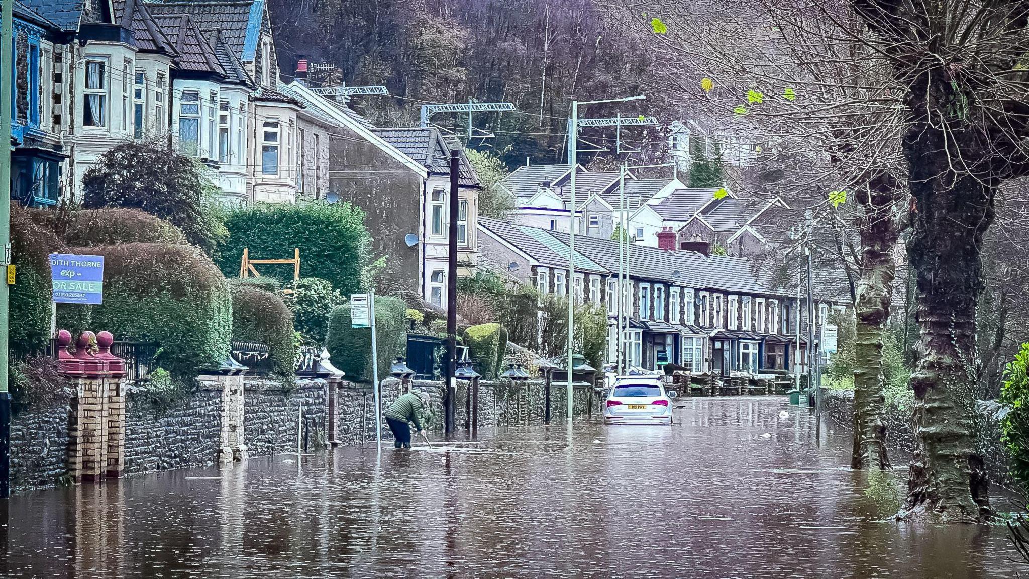 Berw Road in Pontypridd with  the road underwater, and a car up to its wheel well. A person can also be seen standing almost up to their knees in water. 