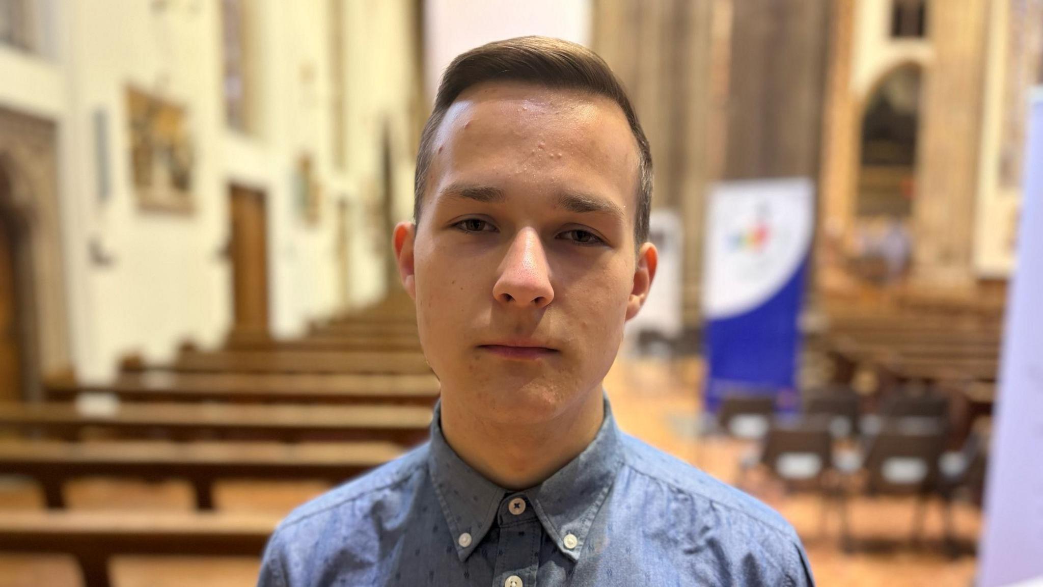 A man wearing a blue shirt looks at the camera while standing in a church hall.
