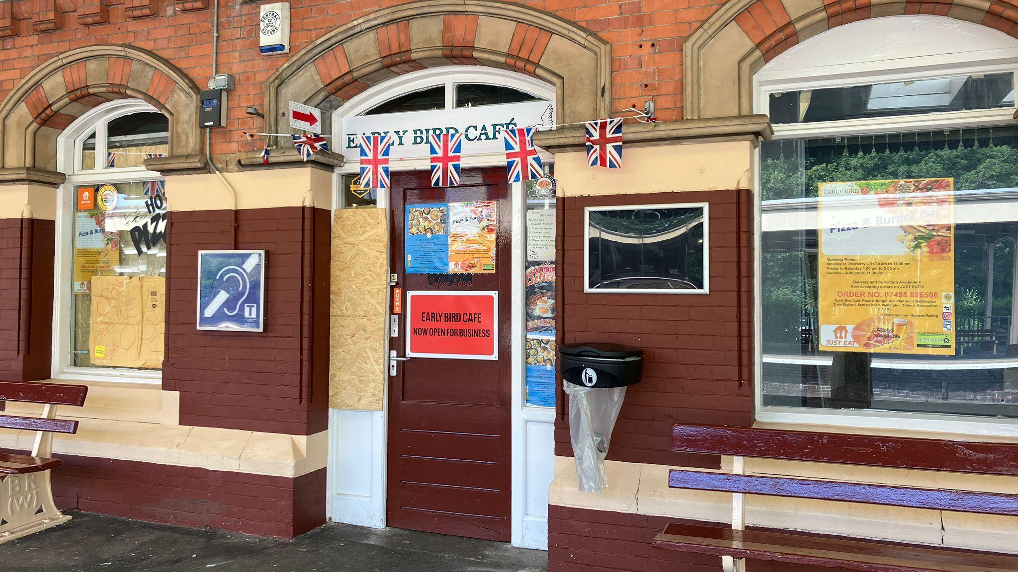 A cafe at a railway station with boarded up windows. The sign above the door says 'Early Bird Cafe'and Union Jack bunting is hung above the door.