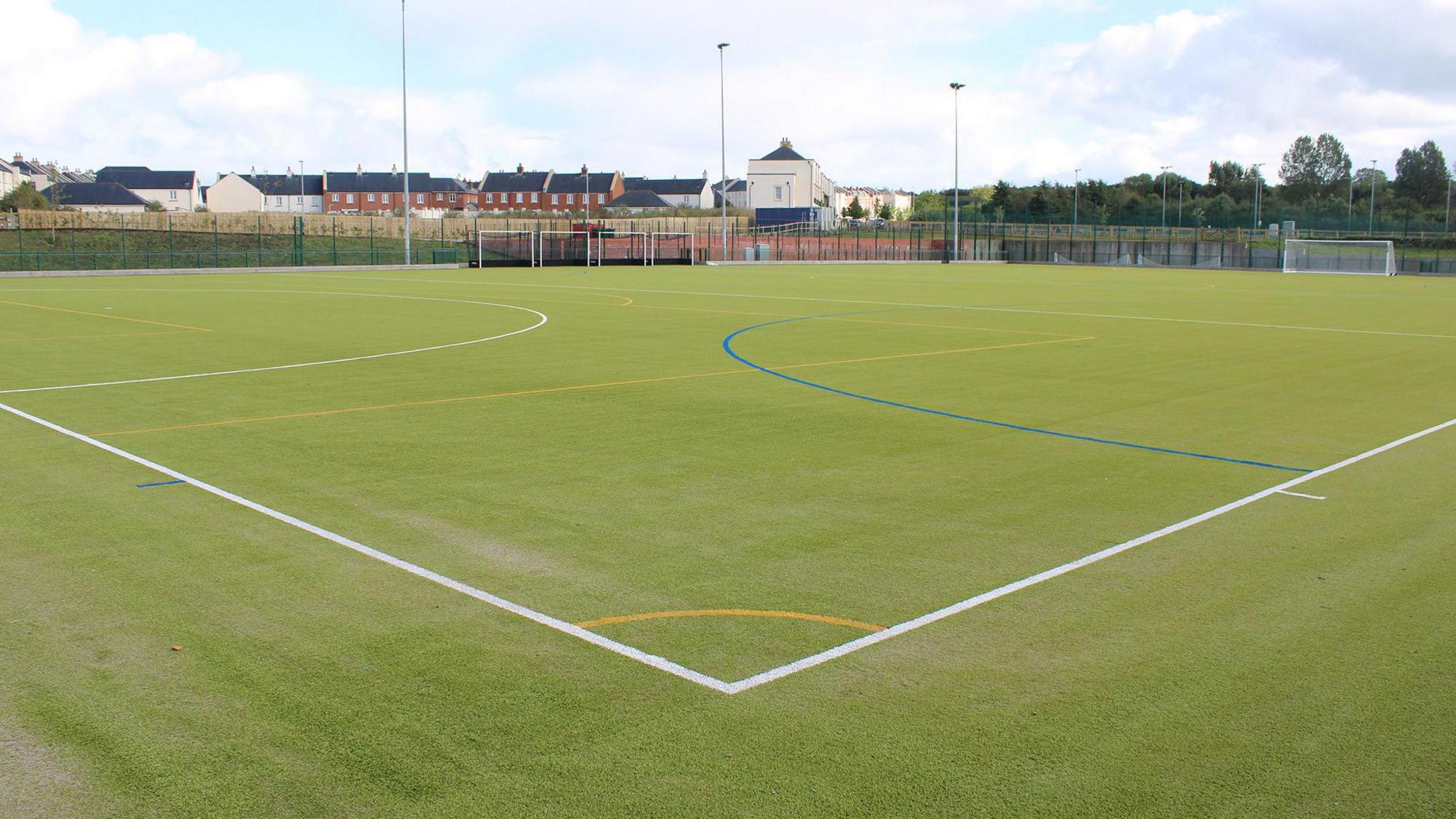 An astro-turf football pitch with a series of lines to mark out the corners and penalty areas and the tops of some red brick houses in the distance.