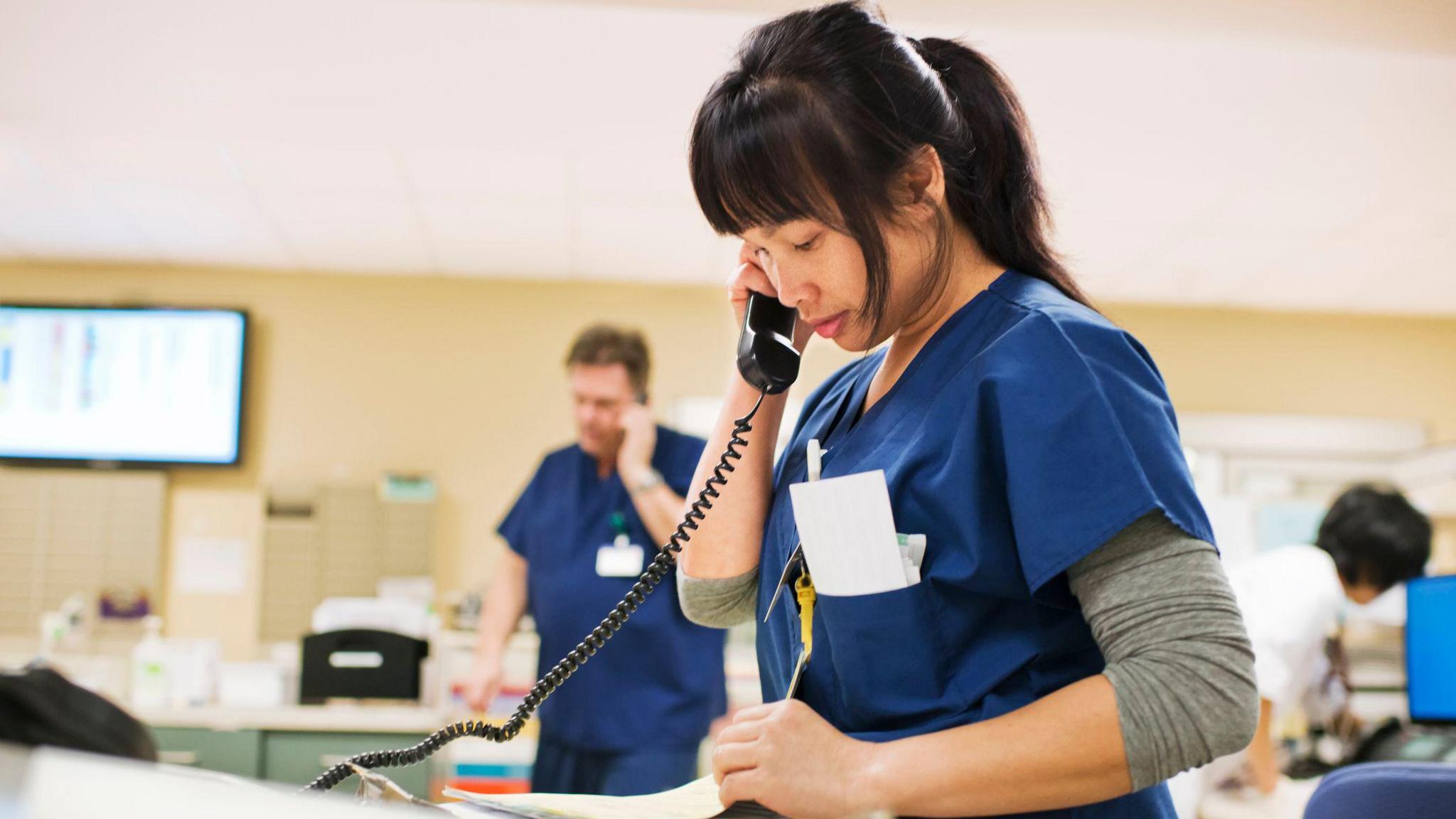 Nurse in blue scrubs takes notes while on the phone