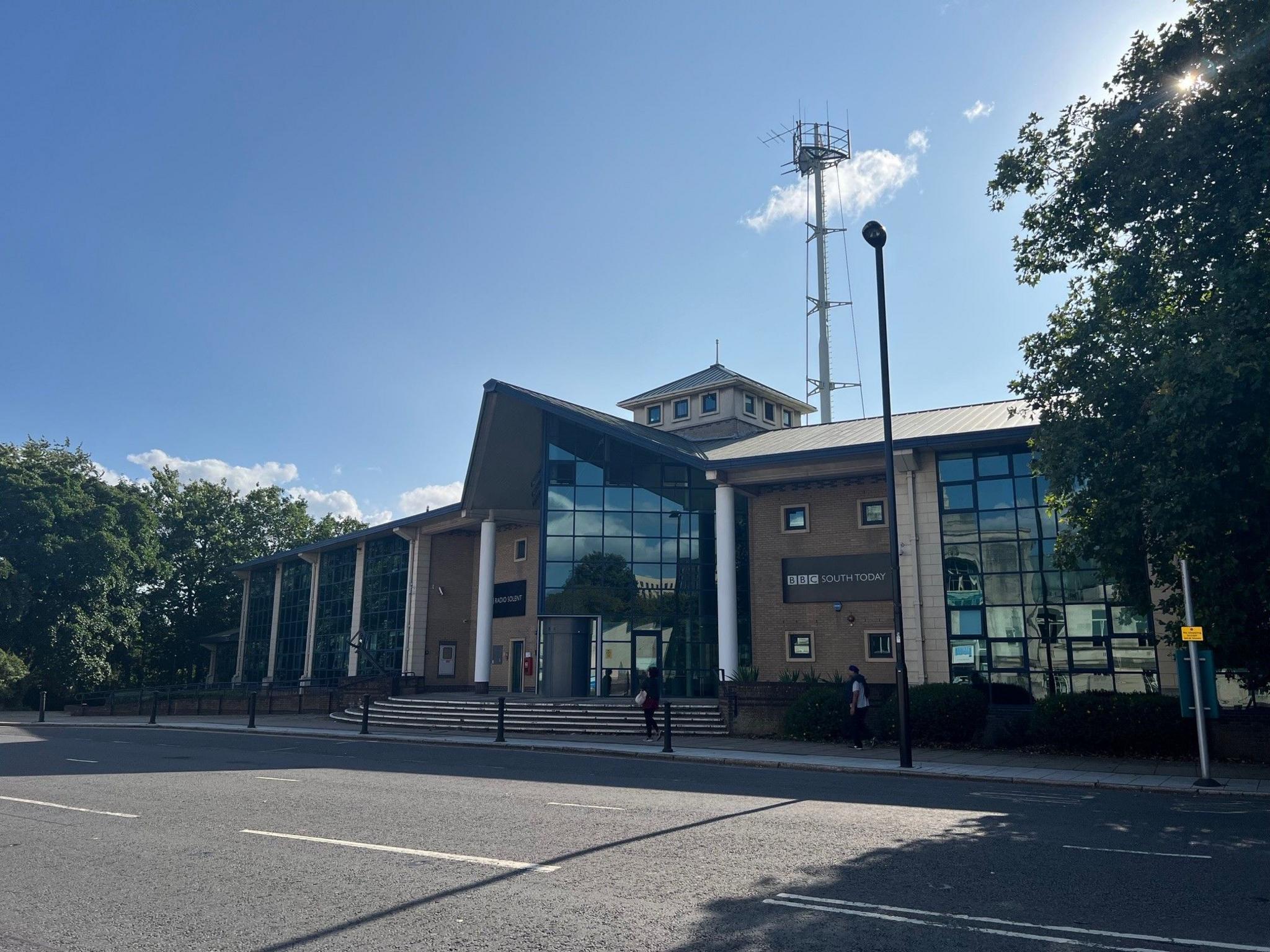 The BBC South building in Southampton. It has a triangular glass facade and a large antenna on the roof. Above it is a bright blue sky
