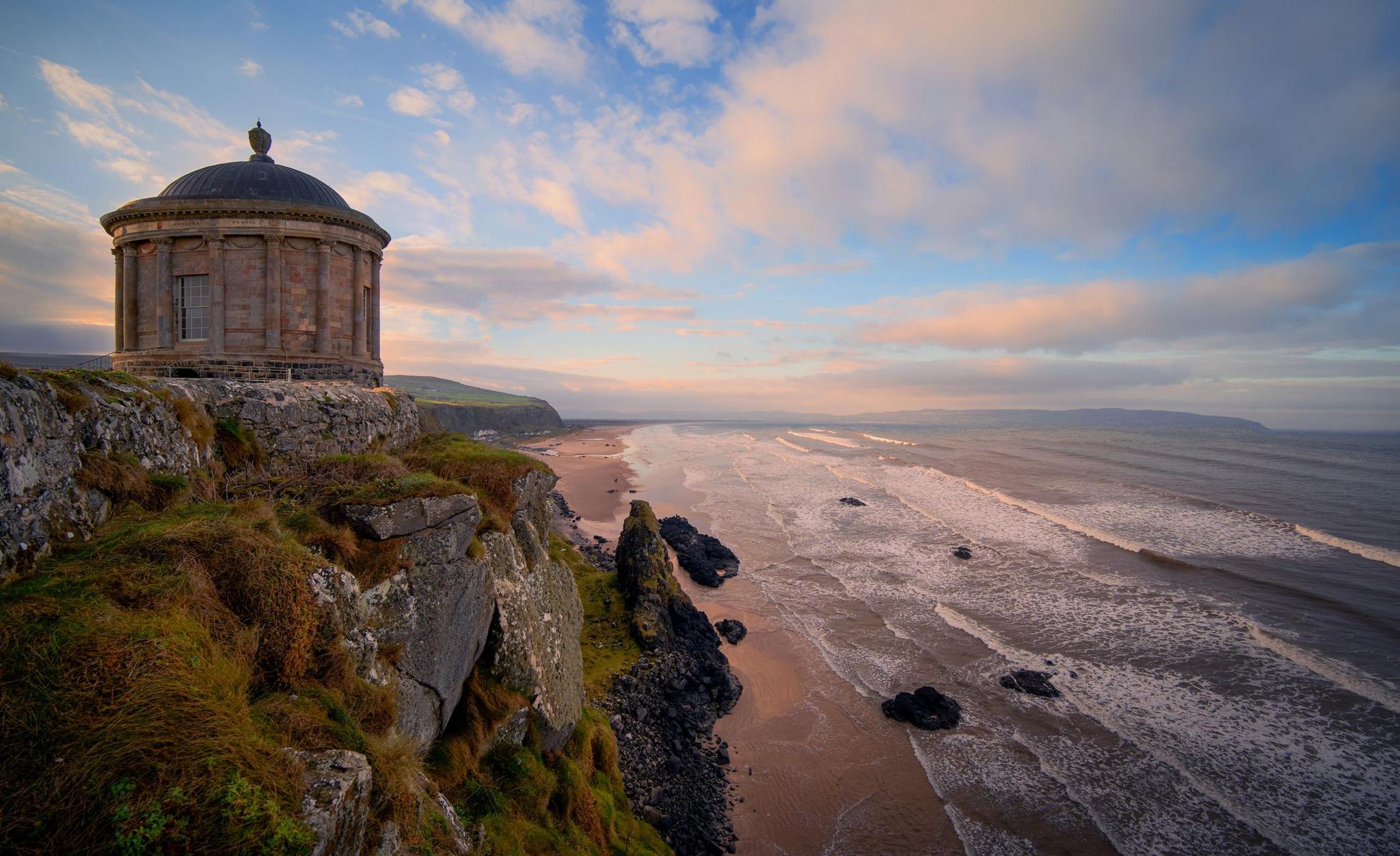 An old temple sits at the edge of a grassy cliff. Below is a beach