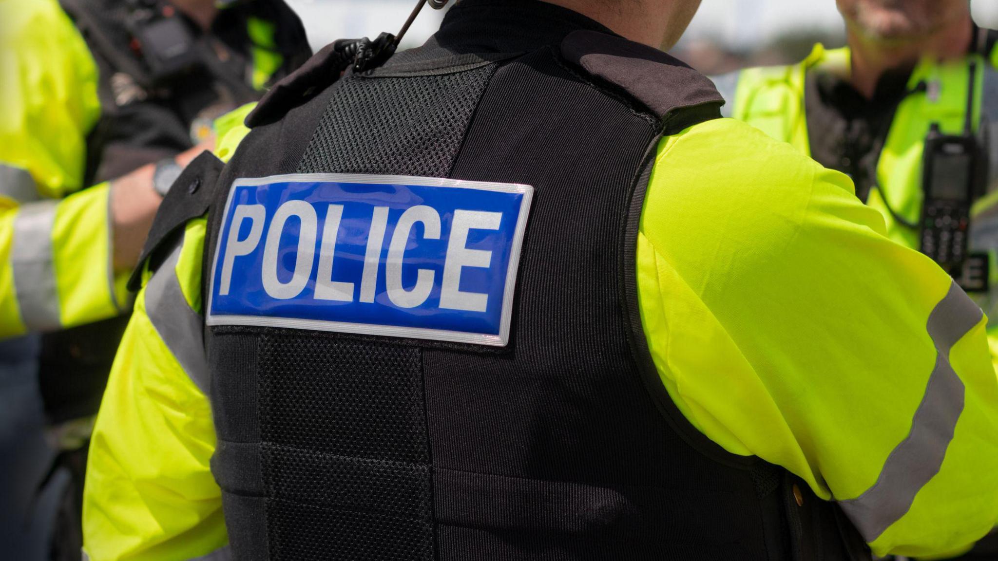 Close-up of 'POLICE' marking written on the back of a hi-visibility stab proof vest worn by a trio of police officers at the scene of an incident. 