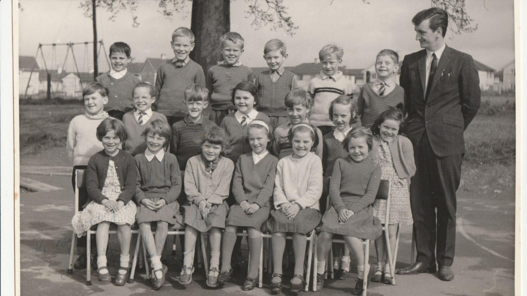 A black and white photo of 19 children sitting on three rows of chairs outside for a school photo, with their teacher in a black suit standing next to them.