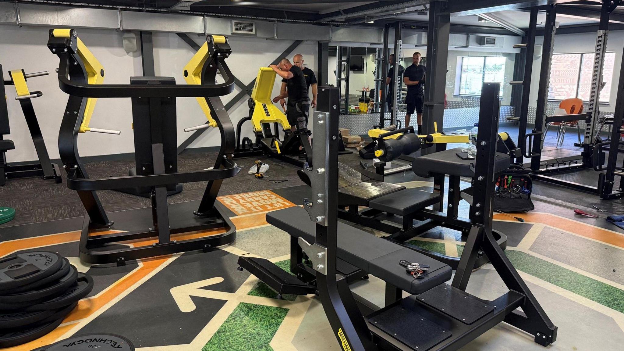 An array of weight machines in a gym. The machines are black and yellow. Some are being assembled by men. There are a mirrors on the wall and tools on the floor.