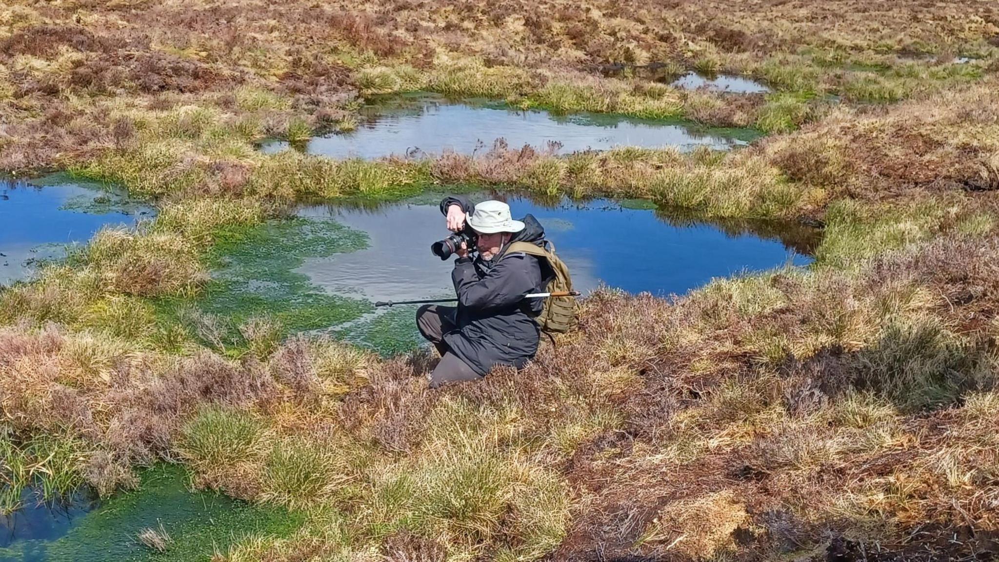 A single person kneels on uneven, grassy terrain surrounded by shallow pools of still water. The person is dressed in outdoor clothing, including a hat and backpack, and is holding a camera up to their face, aiming toward the ground or nearby water. The landscape is open and expansive, covered with brown and green vegetation typical of moorland. Small reflective pools of water are scattered across the scene, some edged with bright green algae. In the background, rolling hills stretch across the horizon under a sky filled with soft, layered clouds.
